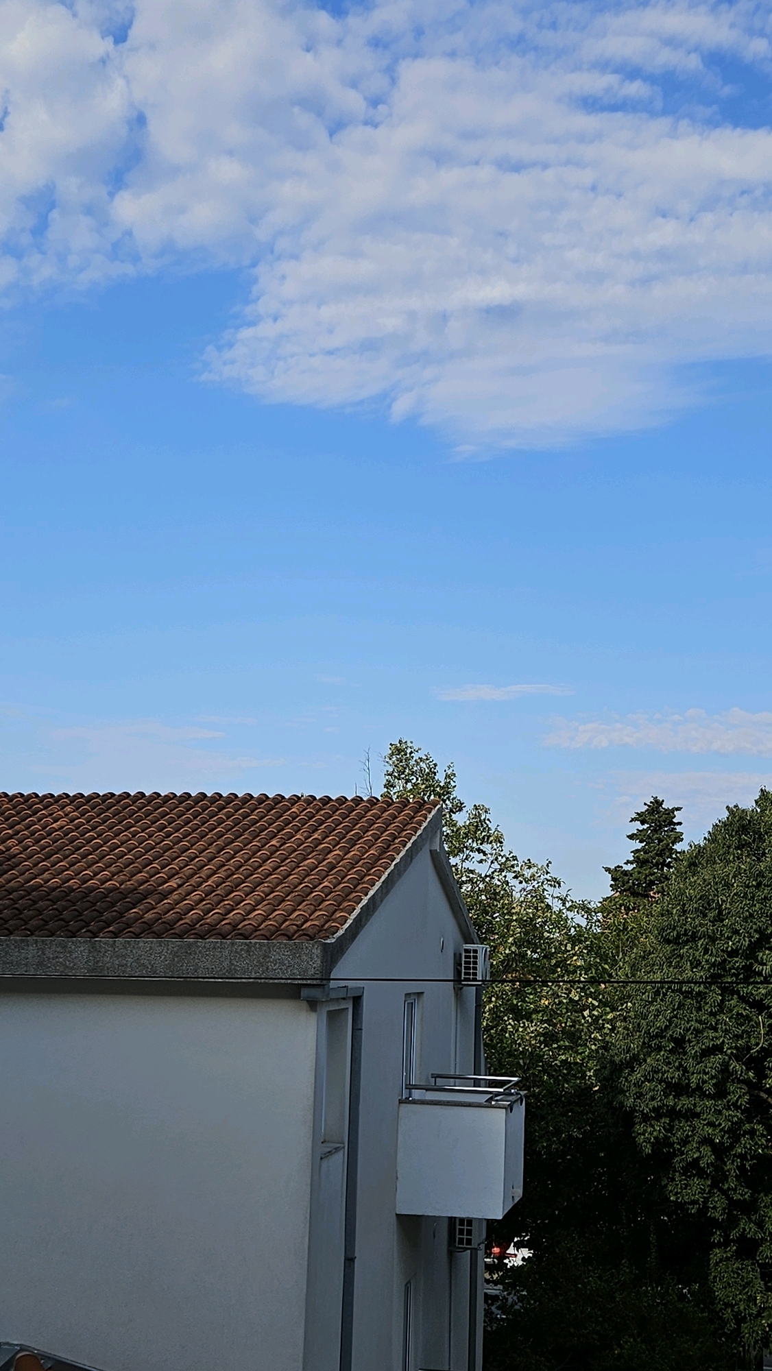 The image shows a portion of a white building with a terracotta tiled roof under a bright blue sky with scattered white clouds. On the side of the building, there is a balcony with white railings and a white facade. An air conditioning unit is visible on the exterior wall near the roofline, and another similar unit is visible lower down. Lush green trees with dense foliage are located behind the building, with some of their branches extending into the frame. A thin black wire runs horizontally across the upper part of the building and tree line. The scene appears to be a residential area in Općina Tučepi, Croatia.