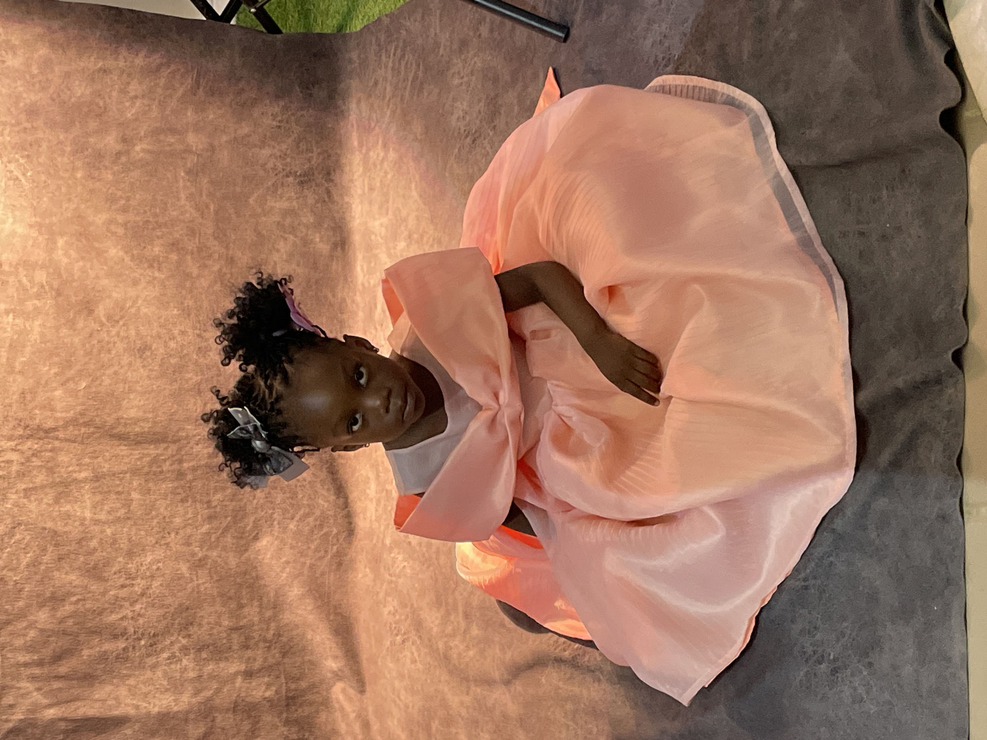 The image is oriented in landscape, appearing to be a portrait photograph rotated 90 degrees counter-clockwise. It captures a young Black girl positioned in the center-left. She has dark, curly hair styled into two buns, each adorned with a gray and white patterned bow. She wears a voluminous, light pink dress made of a shiny, possibly organza-like fabric, featuring a large, elaborate bow detail on the front. Her right arm is visible, resting on the dress. She looks directly towards the viewer with a neutral to slightly curious expression.
The girl is situated on a textured dark gray surface, which serves as the foreground and extends to the right. Behind her is a light brown or tan backdrop with a mottled, textured appearance, suggesting wrinkled fabric or paper. In the upper right corner (relative to the rotated image), a small section of green fabric or turf and a black pole or stand are visible, indicating an indoor studio or photography setup. The lighting is soft, creating highlights on the dress and subtle shadows across the textured backdrops. No text is visible in the image.
