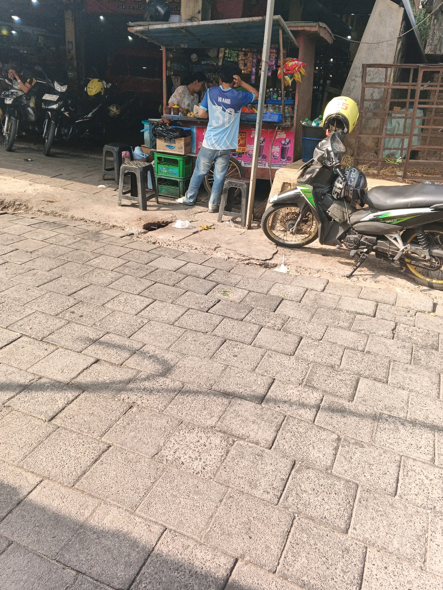 The image captures a street scene in Ungaran, Indonesia, likely near a food stall or small shop. In the background, there's a stall with a canopy, selling various items like drinks and snacks. Two individuals are present near the stall: one person is partially visible, appearing to be an attendant, while another person, wearing a blue and white jersey with the number "30" and the name "WAHID" on it, is standing and looking to the left. Several motorcycles are parked in front of what appears to be a row of shops. One motorcycle in the foreground, a black Honda, is equipped with a yellow helmet on its handlebars. The ground is paved with interlocking stone tiles, and shadows from an overhead structure are cast across the pavement, indicating it's daytime.