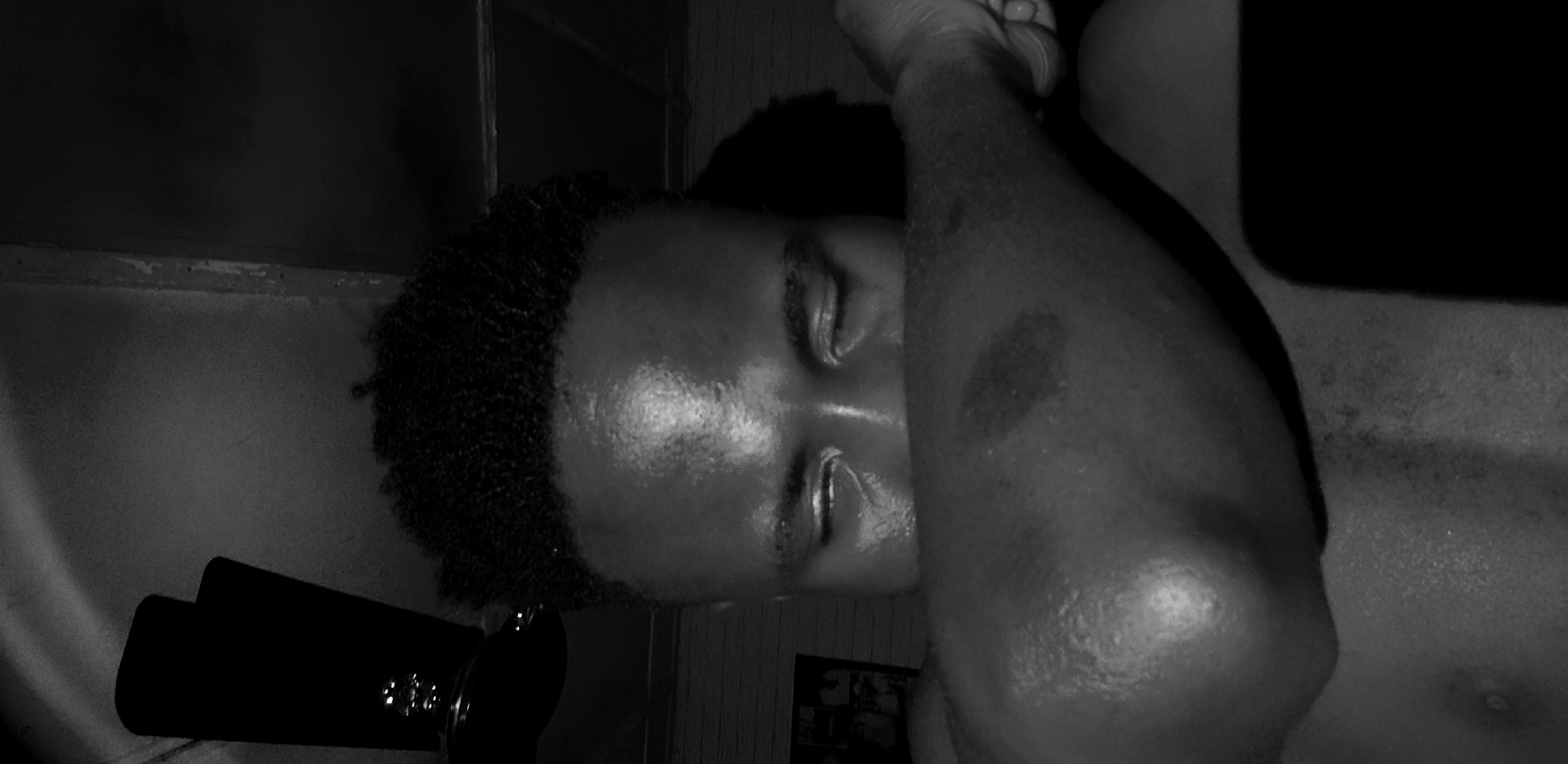 This is a close-up, black and white, low-angle shot of a Black individual, likely male, resting their head on their bicep. The individual's eyes are closed, and sweat glistens on their forehead and neck, suggesting a warm environment. Their hair is short and textured. Part of their arm and shoulder are visible, showing some markings. In the background, a ceiling fan is partially visible on the left, and a brick wall is suggested. The overall lighting is dim, creating strong shadows, particularly on the right side of the frame where a dark, rectangular object, possibly a phone, is present. The image appears to be taken indoors in Narayi, Nigeria.