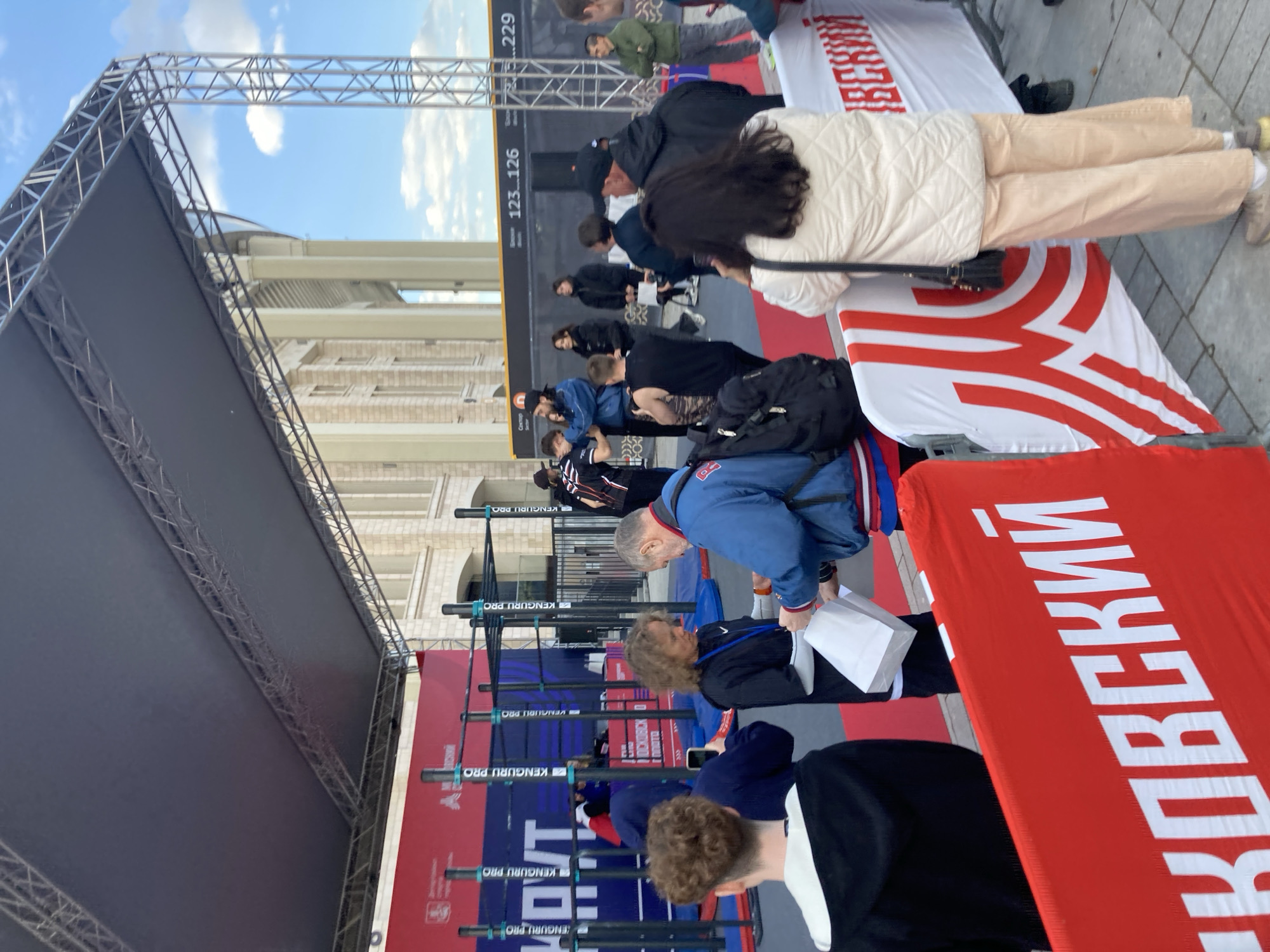 Here is a description of the image:
The image shows a gathering of people in what appears to be a public event or competition. The setting seems to be outdoors, possibly in a city square, indicated by the surrounding buildings and the sky overhead. Several people are standing around a structure equipped with parallel bars, labeled "KENGURU PRO." Red and white banners are present, with Cyrillic text visible, including the word "МOСКОВСКИЙ" (MOSKOVSKIY). A digital display showing the numbers 123-126 and 229 can also be seen, possibly indicating participant numbers or scores. The people present seem to be event attendees or participants, and some are looking at or interacting with the equipment. There's an event canopy overhead. The sky appears to be partly cloudy. The location is likely in Moscow, Russia, due to the Cyrillic writing on the banners.