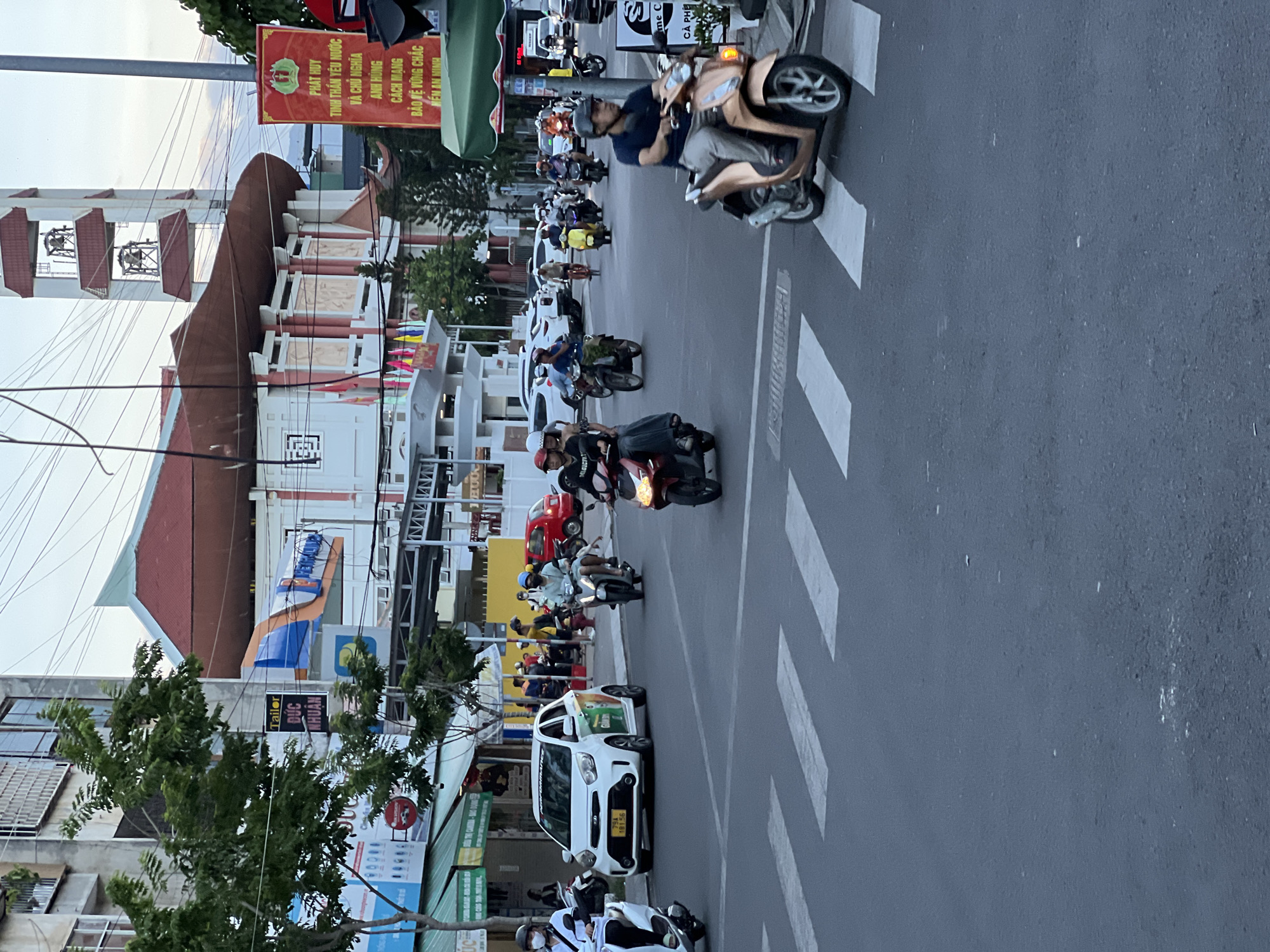 The image captures a street scene in Nha Trang, Vietnam, likely during the late afternoon or early evening. The perspective is from a slightly elevated position, looking down a moderately busy street. A crosswalk is prominent in the foreground, with several motorcycles and scooters in motion and stopped at the intersection.
In the mid-ground, a white Kia Morning car with a license plate "79A 181.56" is parked on the right side of the road. Adjacent to it, on the sidewalk, are several shops with signage, including one with the word "TAILOR" and Vietnamese text. Further down the street, a red car is visible.
The background features a large, modern building with a distinctive red-tiled roof and a bell tower. A red banner with Vietnamese text and an emblem is displayed on a pole to the right of the building. The sky is overcast, suggesting a late day. People are mostly dressed in casual attire, with many wearing masks. The overall atmosphere is one of daily urban life, with a significant presence of motorbikes, characteristic of Vietnamese streetscapes. The architecture suggests a blend of modern and traditional elements.