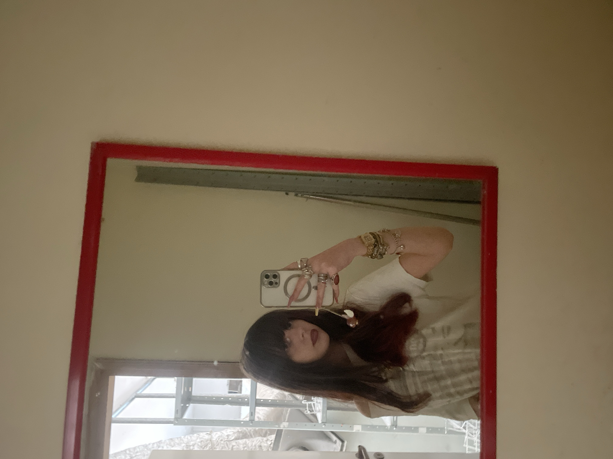 A medium shot depicts a reflection in a red-framed mirror mounted on a cream-colored wall. The reflection shows a person with dark hair and bangs, holding a smartphone in their right hand as if taking a selfie. The person is wearing multiple rings and bracelets. The background visible in the mirror shows a window or opening with a metal structure behind it, suggestive of an interior space. The location in the city of Raanana, Israel cannot be determined from this single image.