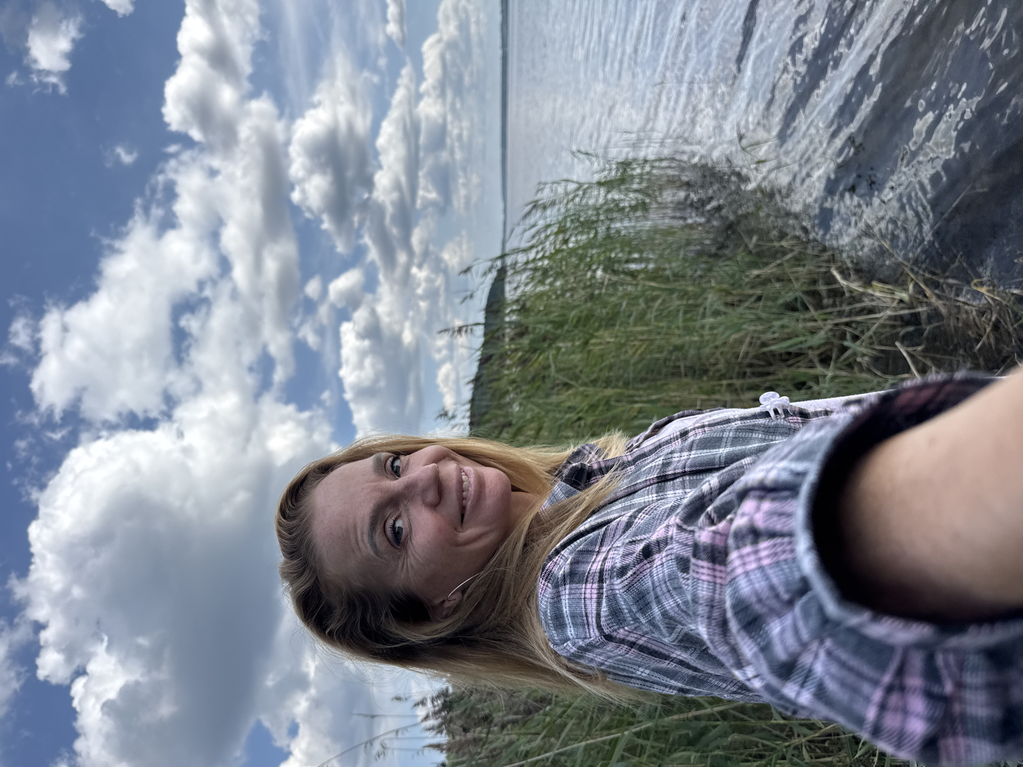 A woman with blonde hair and blue eyes, wearing a pink and grey plaid shirt, smiles at the camera while standing next to a body of water in Portovoe, Russia. She is positioned on the left side of the frame, with the water on the right. Tall, green reeds line the edge of the water, obscuring the ground. The sky above is filled with fluffy white clouds against a bright blue backdrop. The water surface has ripples, suggesting a light breeze. The woman is holding the camera, indicating a selfie.