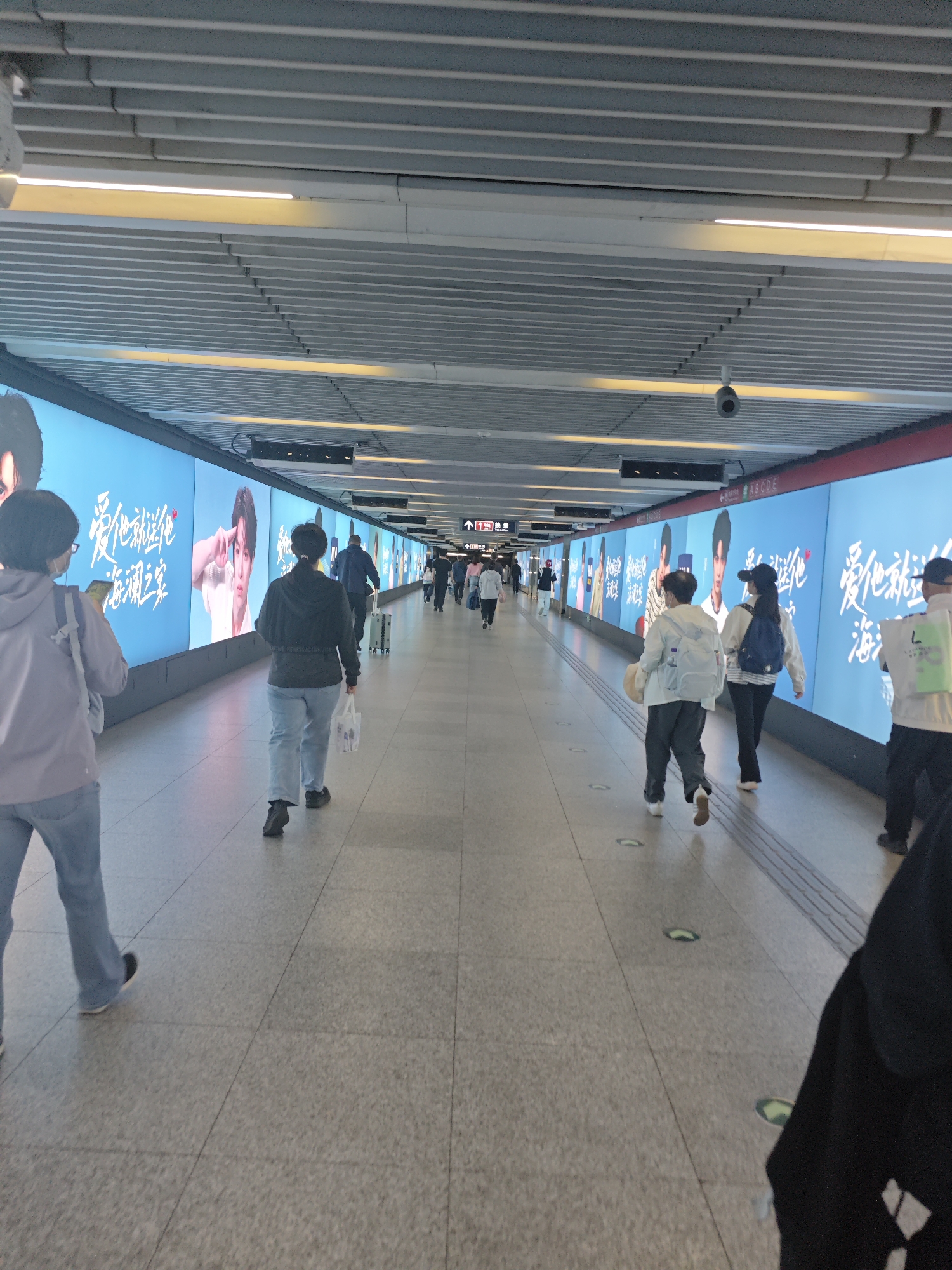 The image depicts a brightly lit, long indoor pedestrian tunnel or hallway, likely within a subway station, airport, or similar public transportation hub. The perspective is from behind a person walking, looking down the length of the tunnel.
Numerous individuals are seen walking in both directions, though predominantly away from the camera. They are dressed in casual attire, with some carrying backpacks or a rolling suitcase. The floor is made of light-colored, square tiled pavement.
The most prominent features of the tunnel are the large, illuminated advertising billboards lining both walls. These billboards display images of Asian individuals, primarily a young man, along with text in Chinese characters. A frequently repeated phrase visible on the billboards is "爱他就是他" (ài tā jiù shì tā), which translates to "Love him, he is him." Another partial phrase seen is "海潮..." (hǎi cháo...).
The ceiling features a slatted design with recessed linear lights providing ample illumination. Several security cameras are mounted on the ceiling, monitoring the corridor. Overhead signs in the distance include an arrow and some unreadable text, as well as a sign displaying "ABCDE." The overall scene suggests a routine passage through a modern, bustling urban facility during what appears to be daytime, or at least well-lit indoor conditions.