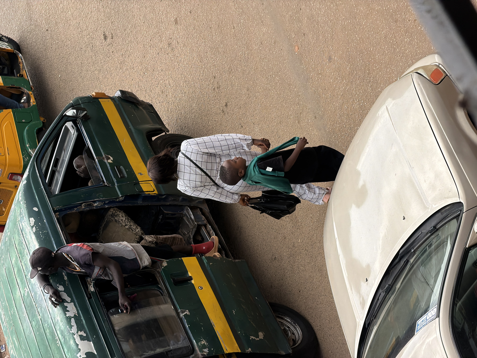 An overhead shot captures a scene on a street in Bukuru, Nigeria. A dark green van with a yellow stripe is parked, its passenger door open. A man wearing a cap and a patterned shirt is leaning out of the open doorway, his arm resting on the van's roof. His left hand is inside the van, and his right hand is visible as he holds a cigarette. A person wearing a checkered white outfit is walking away from the van, carrying a black bag. A young boy in a green sweatshirt over a black t-shirt walks beside them, holding the hand of the person in white. In the foreground, the hood and windshield of a light-colored car are visible. In the background, a yellow and green autorickshaw is parked. The ground is paved with a rough, sandy texture.