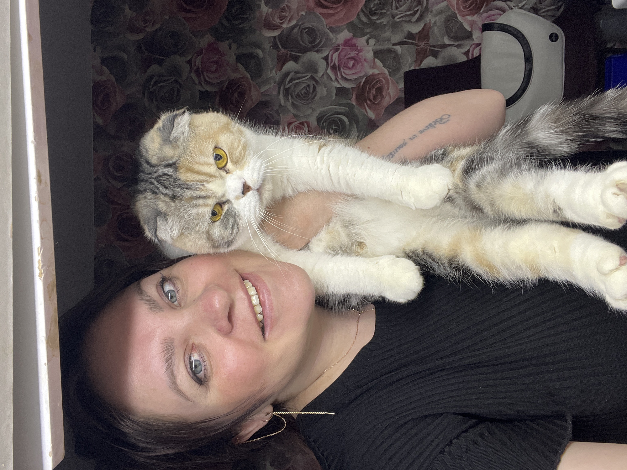 A woman is lying on her back, holding a cat in her arms. The woman has fair skin, blue eyes, and dark hair. She is wearing a black top and long, delicate gold earrings. She is smiling at the camera. The cat is a Scottish Fold with calico coloring. It has yellow eyes and a long, fluffy tail. The background is a floral-patterned wallpaper. There is a white object with a black handle to the right of the woman's arm. A partial view of a window or doorway is visible on the left. The photo appears to be taken indoors. There is no indication of Noviy, Russia.