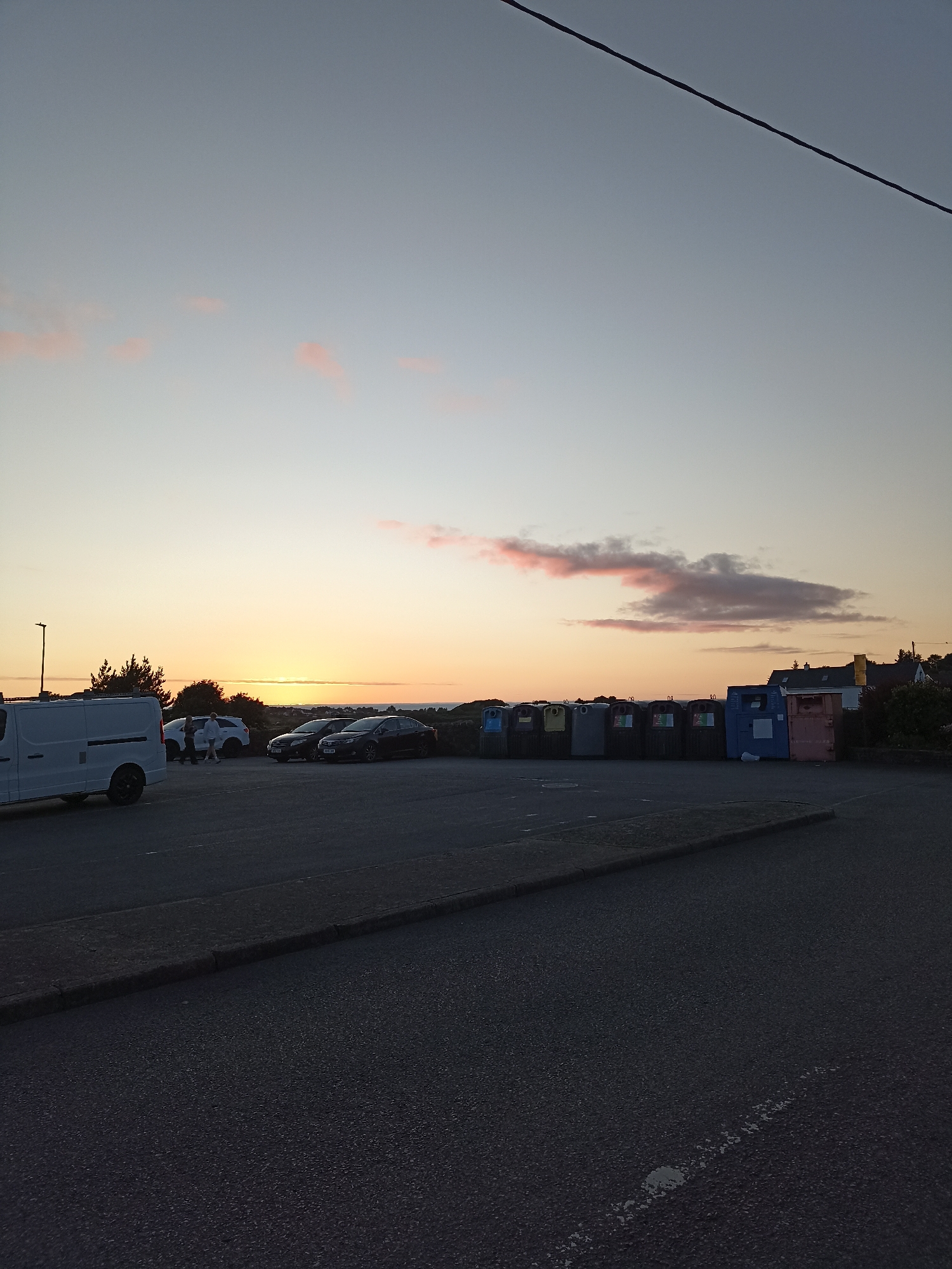 The image depicts an outdoor scene at dusk, possibly in Annagry, Ireland. The sky is a soft gradient of grey and orange, with a few wispy pink clouds. A dark power line stretches diagonally across the upper portion of the frame. In the foreground, a dark asphalt road leads to a parking lot.
Several vehicles are parked in the lot. On the left, a white van is parked. Further into the lot, two white cars and two black cars are visible. Two individuals, seemingly a man and a woman, are standing near the white cars.
A row of large, multi-colored recycling bins (blue, pink, grey, and green) are situated along the edge of the parking lot. Behind the bins, some houses and trees are faintly visible against the twilight sky. The overall impression is a quiet, possibly coastal, location at the end of the day.