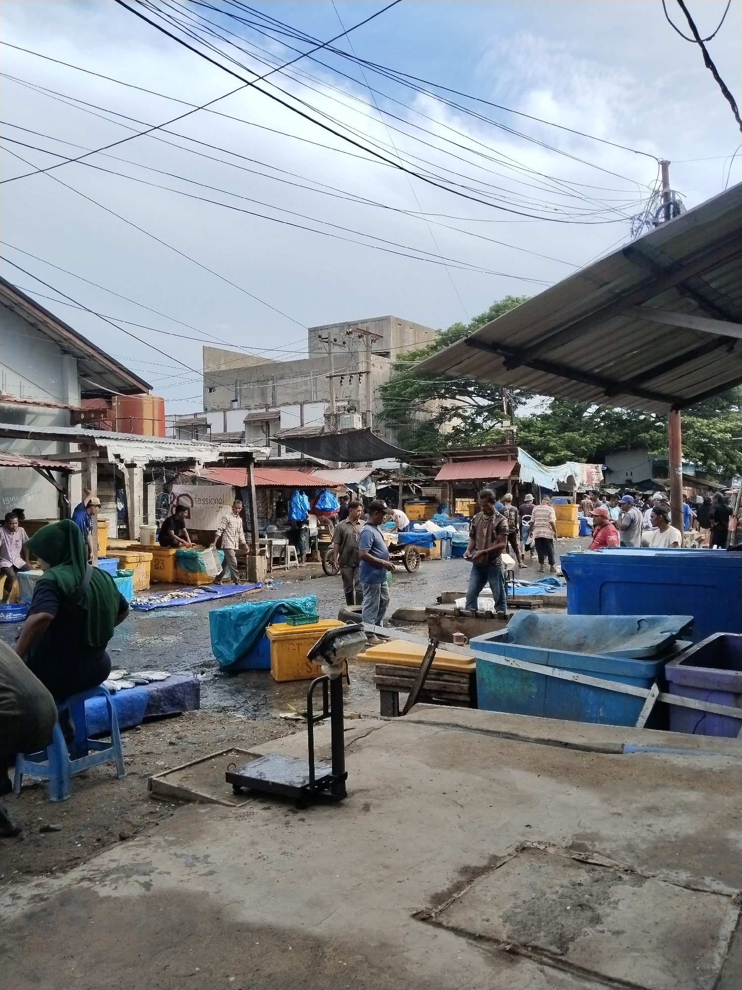 This is a photograph of a bustling fish market scene in Lhokseumawe, Indonesia, under an overcast sky. The market appears to be an open-air setting with various stalls and temporary structures covered by corrugated metal roofs. Numerous people are engaged in activities related to the sale of fish. Several individuals are seen interacting with each other, some appearing to be vendors and others customers. Large blue and yellow plastic containers, commonly used for fish transport, are prominent throughout the scene, some filled with ice and fish. A few of these containers are covered with blue tarps. There are also stacks of wooden crates and what appears to be a traditional scale in the foreground. The ground is wet and appears to be concrete, indicative of a working market environment. In the background, buildings with a mix of modern and older architectural styles are visible, along with electrical wires crisscrossing overhead. The overall impression is one of a lively and active marketplace.