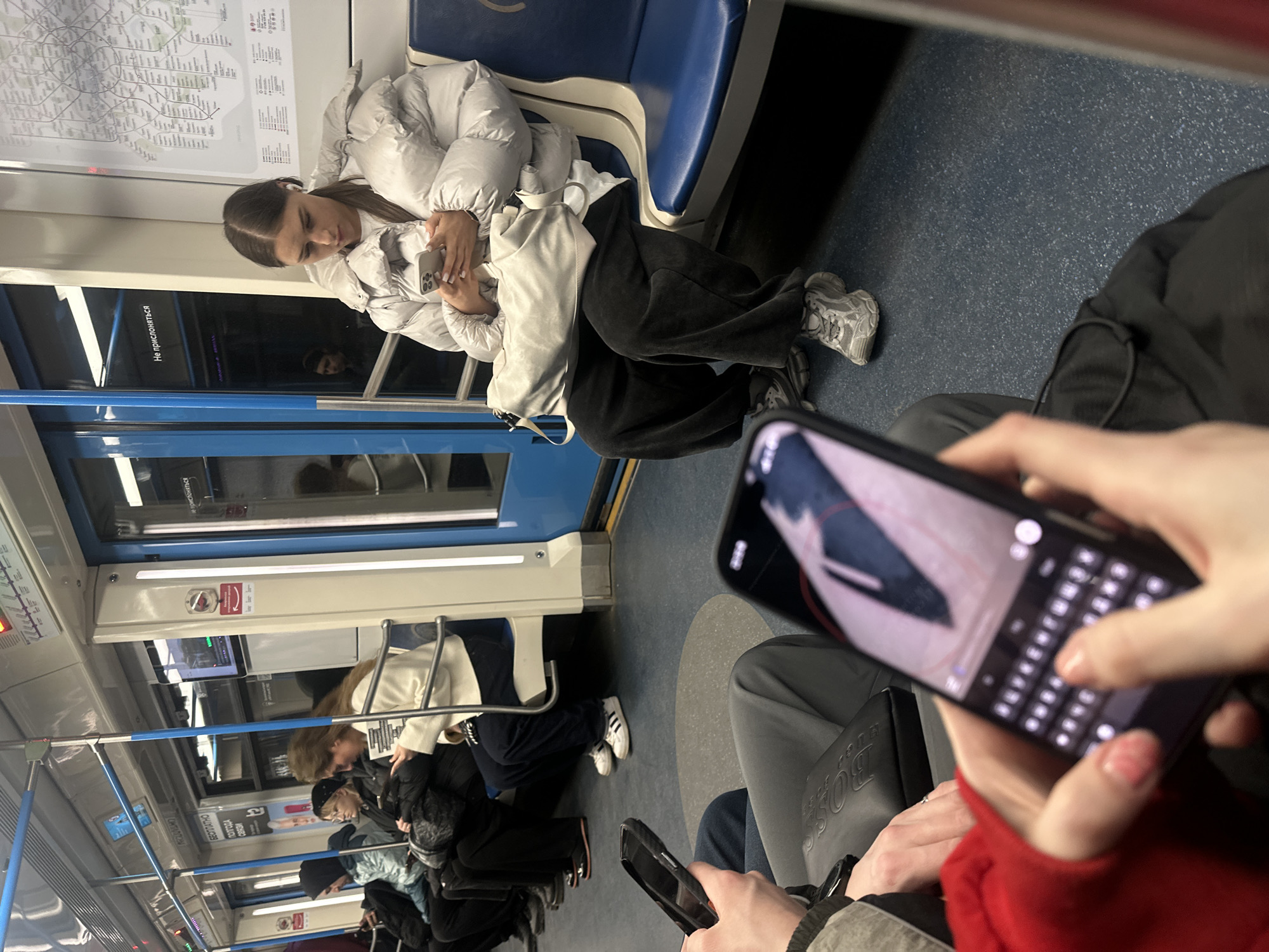 The image shows the interior of a subway car, likely in the city of Dubrovki, Russia. A young woman in a white puffer jacket, black pants, and grey sneakers is seated on a blue subway seat and looking at her smartphone. Other passengers are visible in the background, some seated and some standing. In the foreground, a person is holding a smartphone, with their fingers touching the touchscreen keyboard. A black Hugo Boss bag is near the hand holding the phone.
