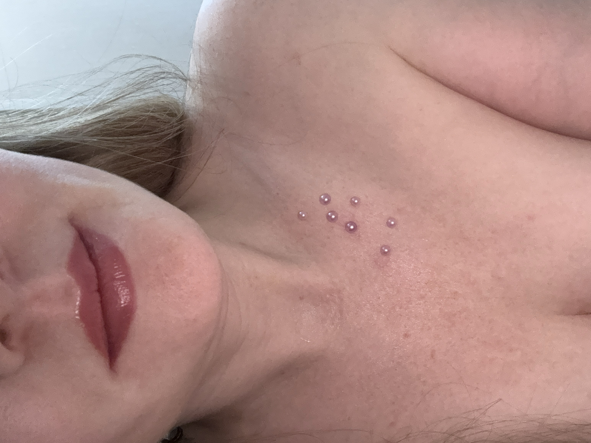 The image presents a close-up view of a person's face, neck, and upper chest area. The subject has light skin and blonde hair. Their lips are coated with a glossy, pink-toned lip product. On the upper chest area, there is a series of small, round, potentially artificial objects attached to the skin. The lighting appears soft and diffused, resulting in minimal shadows and a generally light complexion. The background is a plain, light-colored surface. There is no indication of the city of Chaikovsky, Russia in the image.