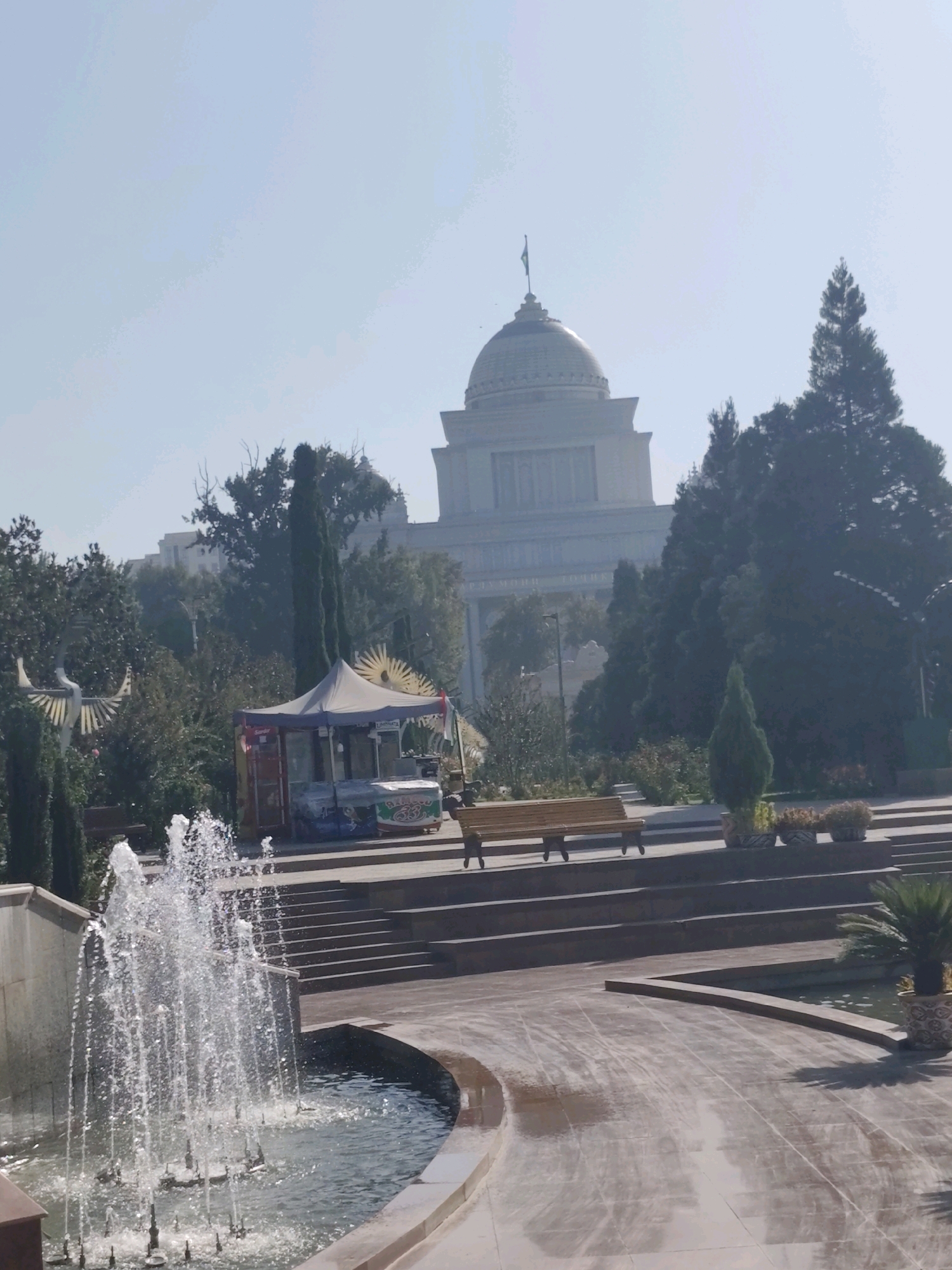 The image depicts a park scene in Dushanbe, Tajikistan, with a prominent domed building in the background, likely a government or cultural institution. In the foreground, a fountain sprays water into a pool, with stone steps leading up to a shaded area featuring a small kiosk or food stand. Several trees, including some tall, slender evergreen trees, are scattered throughout the park. A wooden bench is visible on one of the stone tiers. The overall atmosphere is one of a sunny day in a well-maintained public space.