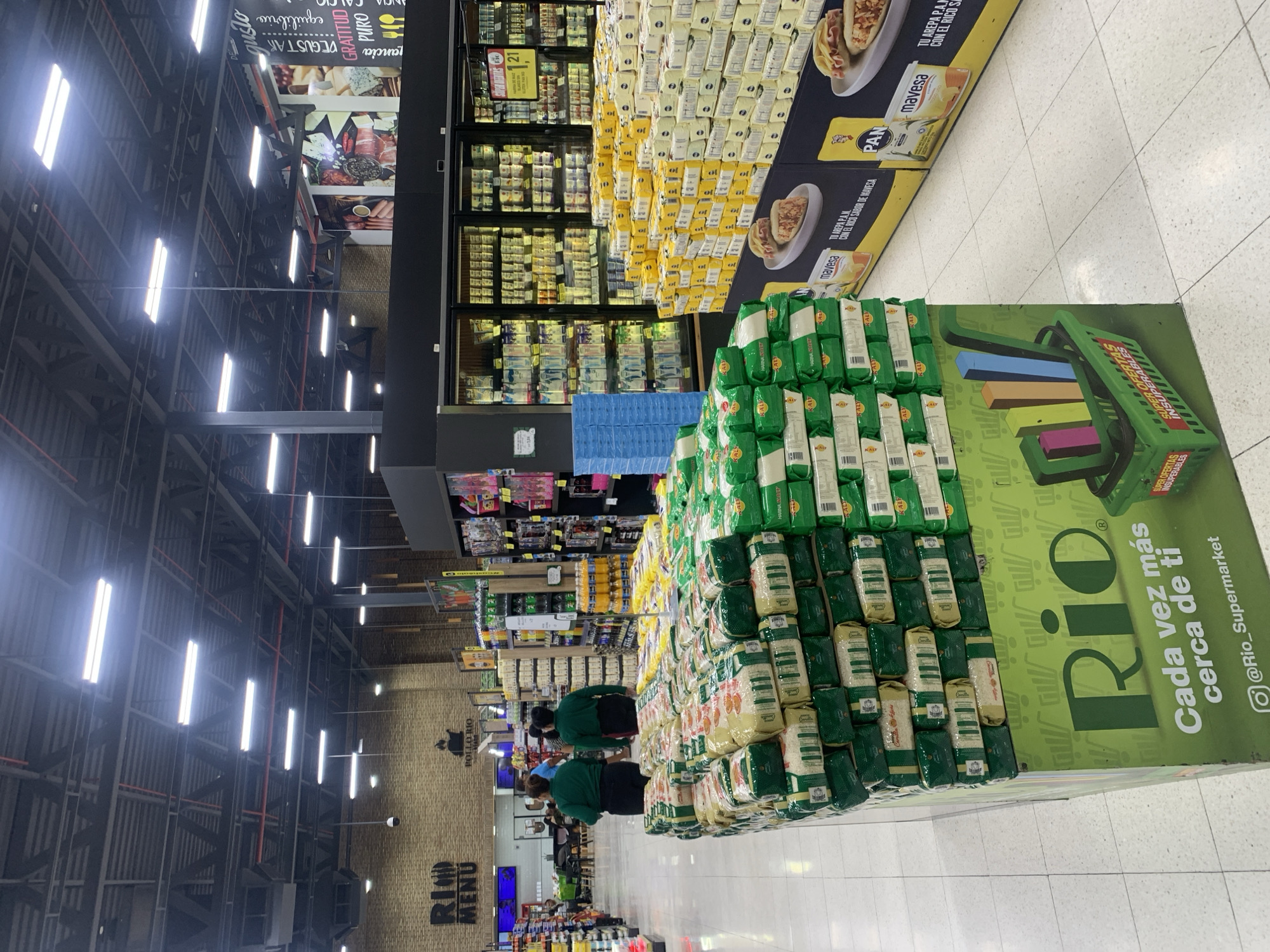 This indoor photograph, rotated to the left, captures a brightly lit supermarket interior in Juan Griego, Venezuela, likely a "RIO Supermarket" as indicated by signage. The scene is dominated by large product displays and high industrial ceilings with rows of fluorescent lighting.
In the foreground and midground, massive stacks of packaged goods are visible. To the right, a large, floor-standing display features numerous green-packaged bags, identifiable as "HARINA KALY" (Kaly flour), arranged in a pyramid shape. Next to it, a green display stand features the "RIO" logo and slogan "Cada vez más cerca de ti" (Closer to you every day), along with "SUPER OFERTAS INSUPERABLES" (Unbeatable Super Offers) and the Instagram handle "@Rio_Supermarket".
Behind these, another prominent display showcases stacks of yellow-packaged bags, likely flour or other staples. On the left side of the image, rows of refrigerated display cases filled with various products line the wall. Adjacent to these cases, cardboard displays for "PAN" brand flour feature images of prepared "arepas" and the text "TU AREPA PAN CON EL RICO SABOR DE MAVESA." Another vertical poster-like display near the ceiling promotes food items with words like "gusto," "GRATITUD," "PURO," and "DEGUSTAR."
A few individuals, possibly employees in matching green shirts or shoppers, are visible further back among the aisles, but their faces are not clearly discernible. The overall setting is that of a bustling, well-stocked retail environment, suggesting a typical daytime shopping experience. The floor is tiled in a light color, and the upper walls are brick, with "POLLO RIO" and "RIND MENU" signs seen on a distant wall.