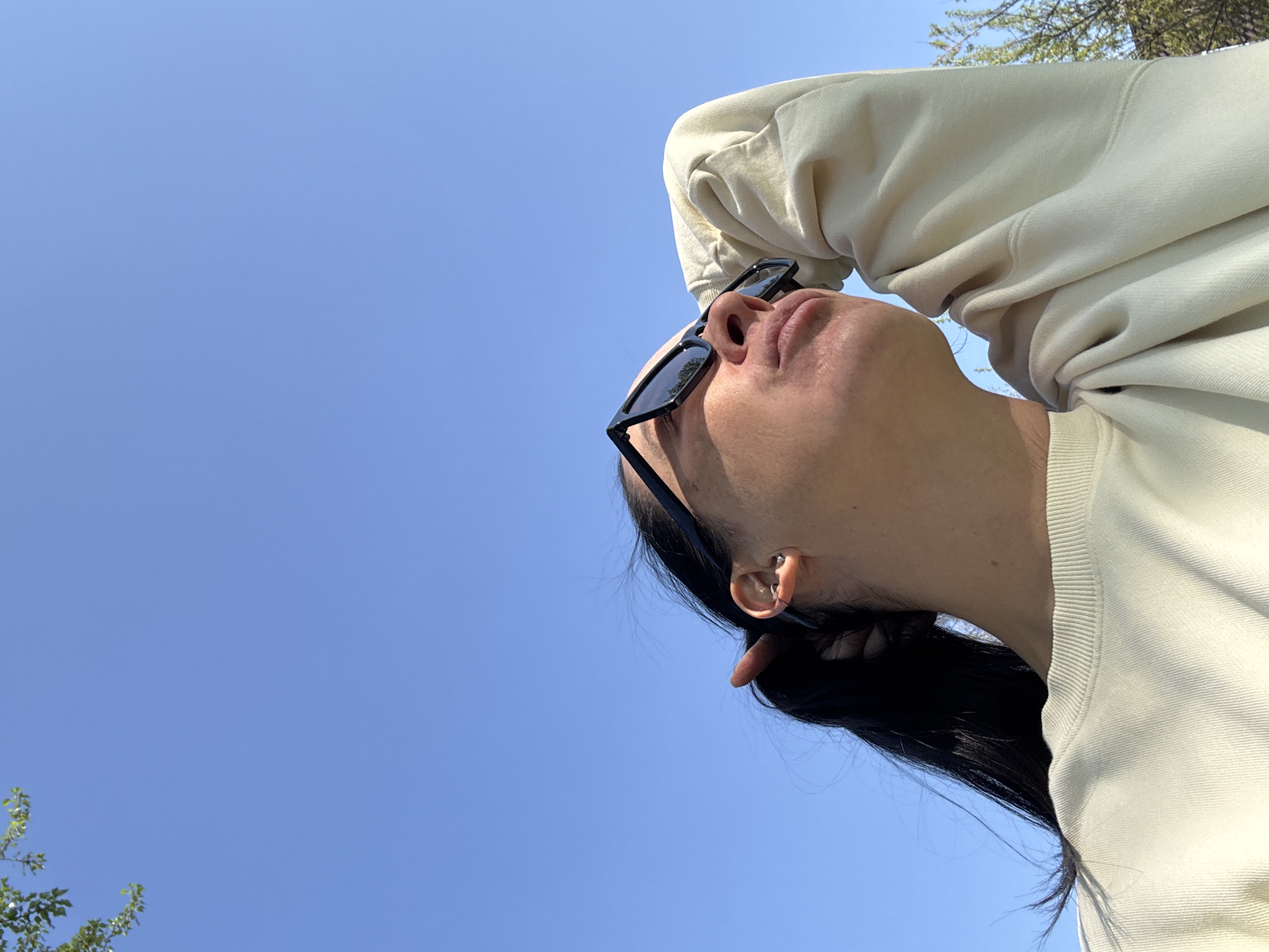 The image shows a person wearing a light-colored sweatshirt and black sunglasses, looking up at the sky. The person has black hair pulled back. The background is a clear blue sky with some tree branches visible in the upper right corner and lower left corner of the frame. It is not possible to determine the specific location in Chaikovsky, Russia from this image alone.