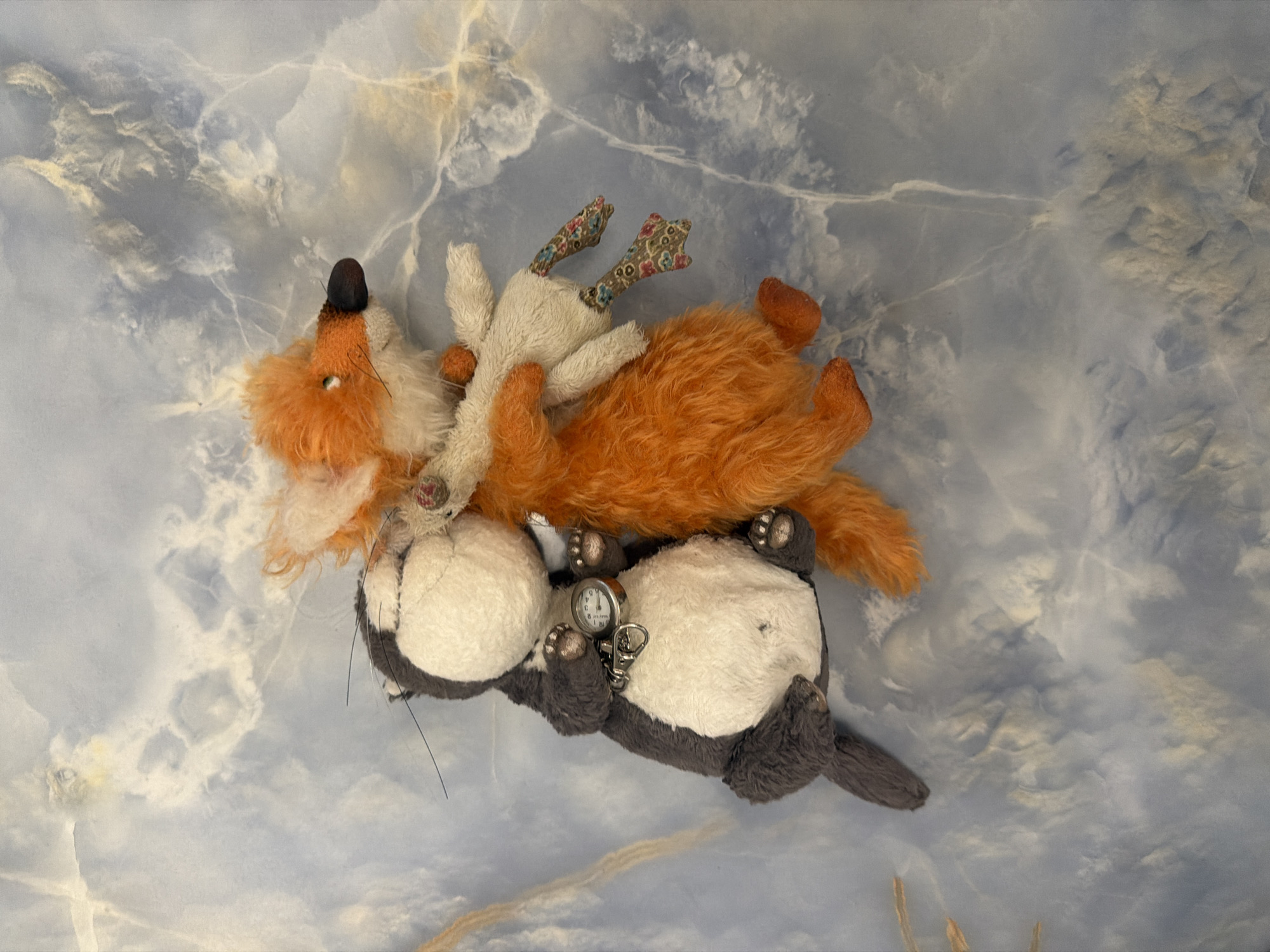 A stuffed grey and white cat is embracing a stuffed orange fox from behind. The cat is on the left, facing right and slightly away from the viewer. It has black whiskers and is wearing a silver chain around its neck with a small silver watch attached. The fox is on the right, facing left and slightly away from the viewer. It has white fur around its muzzle and chest, with orange fur elsewhere. The fox is holding a small stuffed white creature with patterned legs in its paws. The stuffed animals are positioned against a blue and white marbled background. The image is taken from a top-down perspective.