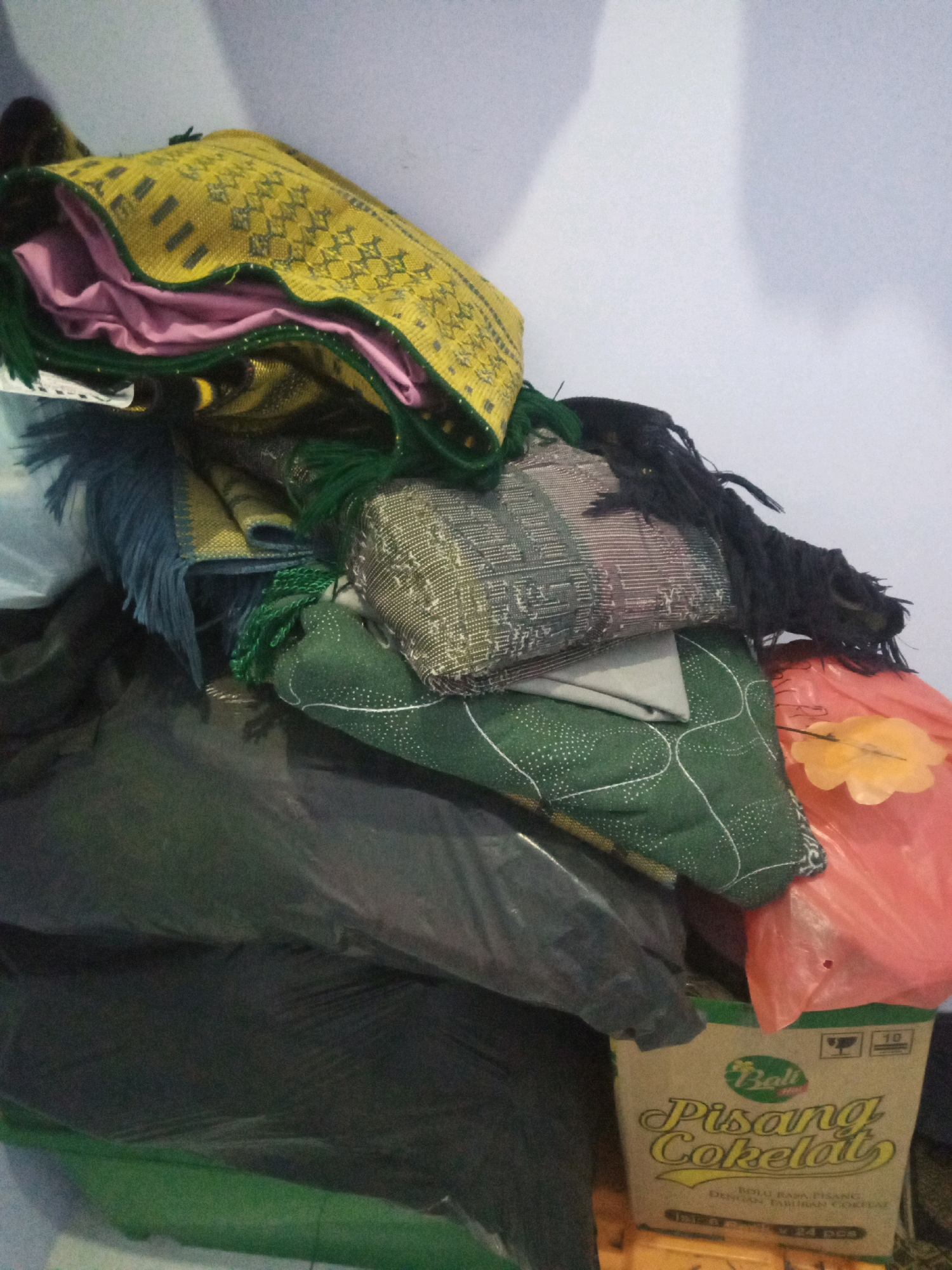 In Sumbergirang, Indonesia, a pile of textiles is stacked against a light blue wall. The textiles consist of several prayer mats, some with intricate geometric patterns in yellow and green, and others in darker tones with fringe. One mat features a pink fabric underneath it. Next to the mats, a pile of green and grey fabrics with embroidered patterns are visible. The lower part of the image shows dark green and black plastic packaging. At the bottom right, a box labeled "Bali Pisang Cokelat" is partially visible, and a translucent orange plastic bag with a yellow flower-like object inside is also present. The scene appears to be indoors, possibly a storage area, with the lighting suggesting a dimly lit environment.