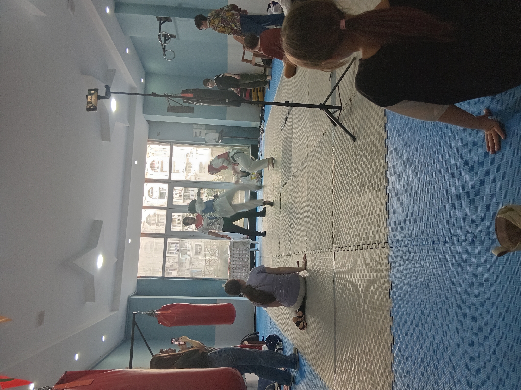 A horizontal indoor shot shows a taekwondo class in progress. In the foreground, a woman with her back to the camera does push-ups, while another woman kneels with her hands on the floor. A tripod holding a smartphone is positioned near the women. In the background, several people are practicing taekwondo, and others are watching. Some individuals wear face masks. Red punching bags hang from the ceiling. Several people are recording the practice with their smartphones. The room has blue and grey puzzle mat flooring. Outside the windows, buildings are visible. The location is Batumi, Georgia.