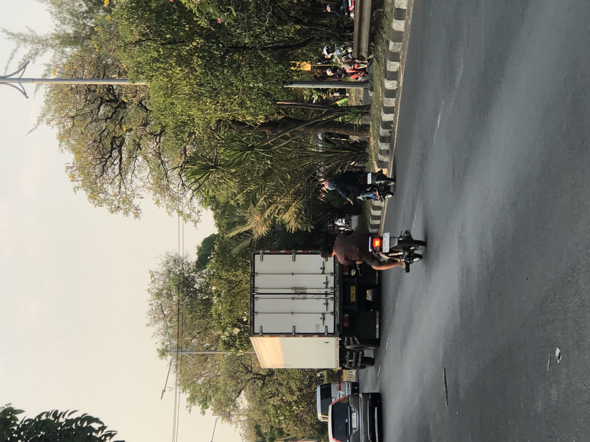 The image captures a street scene in Ngaban, Indonesia, bathed in the soft light of what appears to be late afternoon or early morning. The foreground features a grey asphalt road with some debris scattered on its surface. Several vehicles are present: a grey SUV is visible on the left, a white van is behind it, and a large white box truck is in the center of the frame, moving away from the viewer. A man on a motorcycle, wearing a dark shirt, is in the immediate foreground, also heading away from the camera. To the right of the truck, a group of motorcycles with riders, some wearing helmets, are moving along the road. The background is dominated by lush green trees and foliage lining the street. Streetlights and utility poles are also visible, suggesting an urban or suburban setting. The road has distinct black and white striped curbing along the right side.