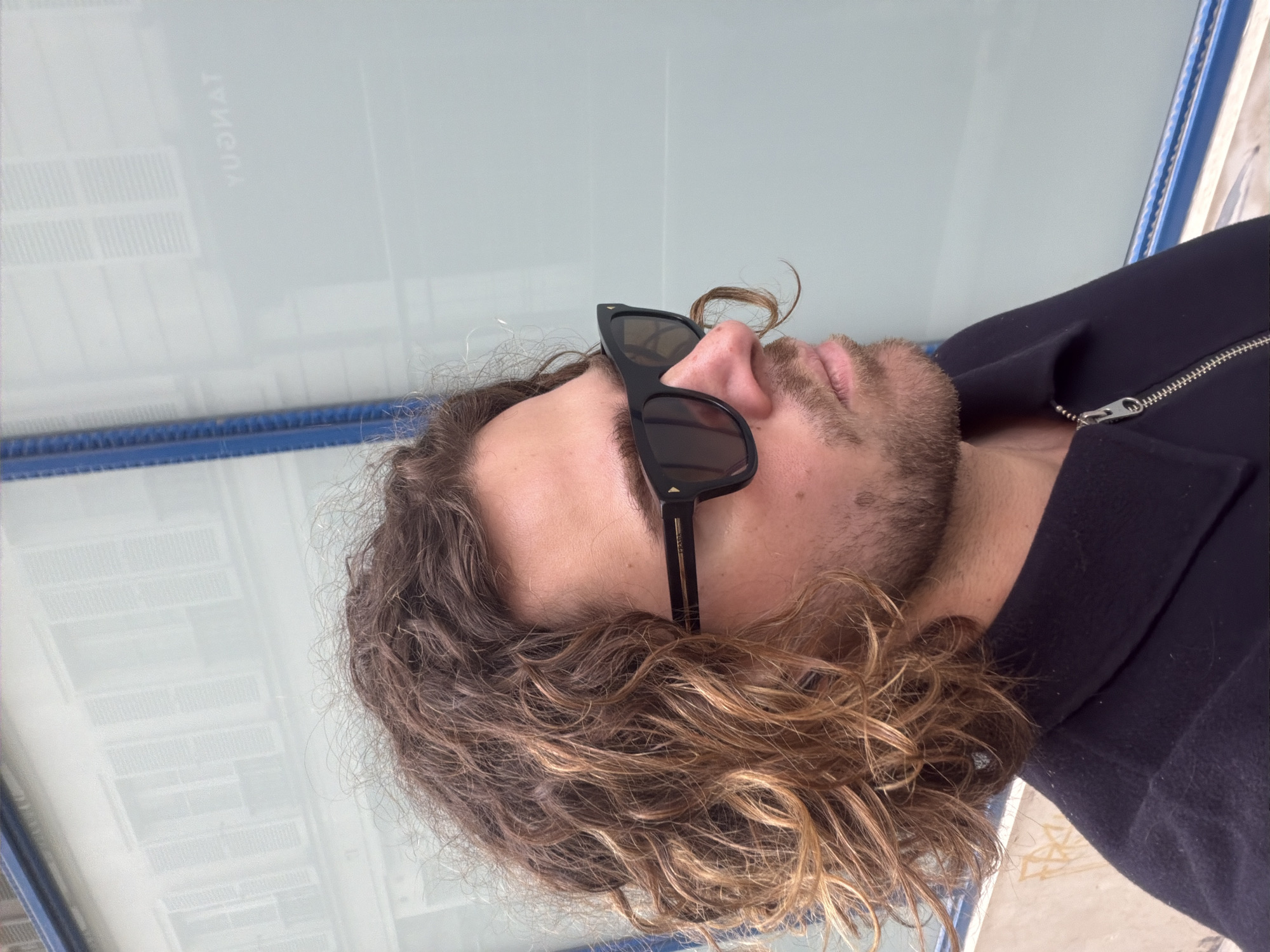 A medium close-up depicts a man with curly brown hair and a beard, wearing black sunglasses and a dark zip-up jacket. He is standing next to a bus stop shelter. The shelter has a blue frame and a glass panel. The word "TANQURY" is visible on the glass. This photograph was taken in Paris, France.