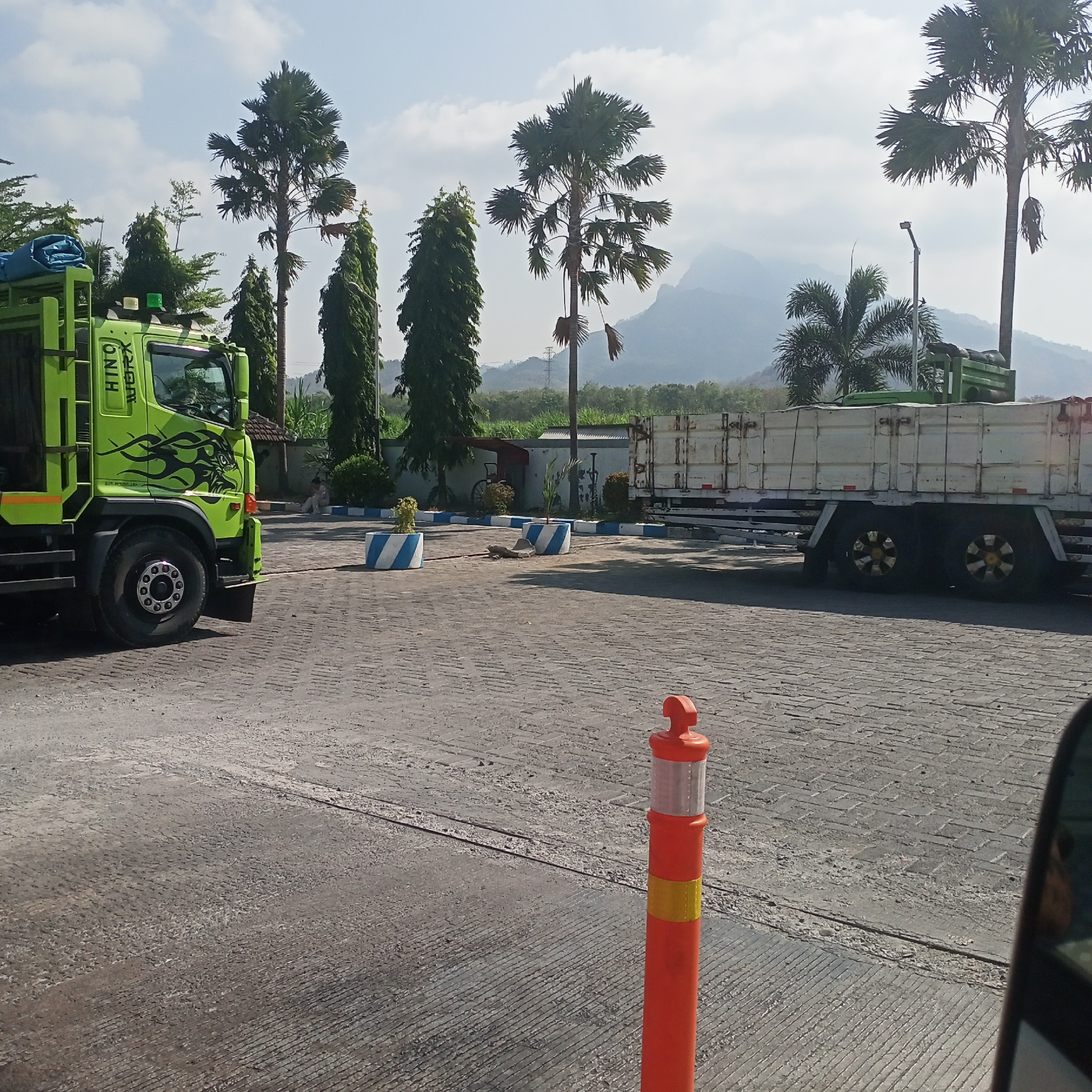 In Klatakan, Indonesia, this image captures an outdoor scene with two trucks, palm trees, and mountains in the background. A bright green HINO truck is prominently featured on the left, with black flame decals on its side and a blue tarp on its bed. To the right, a white trailer truck with dual rear wheels is parked. The ground is paved with interlocking grey bricks, and a row of decorative planters with blue and white stripes are visible. A solitary orange traffic cone stands on the right side of the frame, and the distant landscape is characterized by lush greenery and hazy mountains under a partly cloudy sky. A person is seated on the ground near a building in the middle ground, and other vegetation, including tall, slender trees, is scattered throughout the scene. The overall atmosphere suggests a sunny day in a somewhat industrial or logistical area.