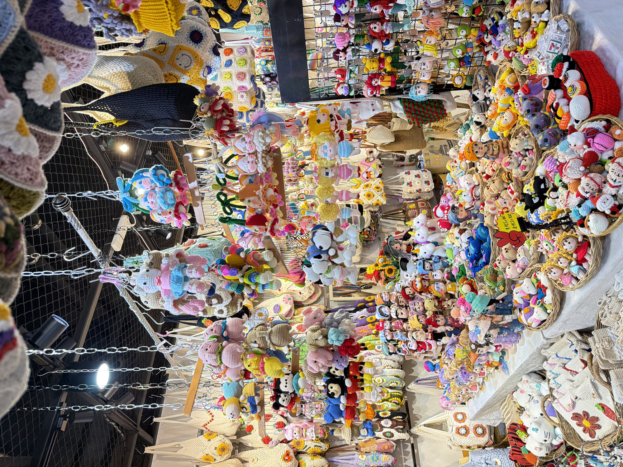This image captures a brightly lit, densely packed market stall specializing in handmade crocheted goods, highly indicative of a vendor at the Chatuchak Weekend Market in Bangkok, Thailand. The ceiling features exposed black track lighting fixtures and fluorescent panels, with numerous metal chains descending to display merchandise.
The stall is overflowing with a vibrant assortment of crocheted items. Hanging from various rods and chains are numerous crocheted bags, many featuring distinct motifs such as sunflowers, smiley faces, cherries, and diverse geometric or floral patterns. Several larger, decorative crocheted pieces, some with flower designs, are also visible hanging from the ceiling.
Below these hangings, the display extends across tables and tiered racks. A multitude of crocheted keychains and small figures are arranged in clusters and baskets. These include popular characters like Hello Kitty, Mickey Mouse, and Minnie Mouse, alongside numerous animal figures such as elephants, bears, unicorns, frogs, and bees. Also prominent are various human-like dolls, mermaids, and even miniature crocheted sushi sets and Santa Claus figures. Price signs are visible, one indicating "1 PCS = 120, 2 PCS = 100" (likely Thai Baht). Some items are packaged, with one showing the "7 ELEVEN" brand logo.
The items are presented in a highly organized but voluminous manner, utilizing woven baskets for smaller figures, wire grid racks for hanging keychains, and display tables for various sized pieces. The overall impression is one of intricate craftsmanship and overwhelming variety, typical of a bustling craft section within a major Southeast Asian market. No individuals are visible interacting with the merchandise in the frame.