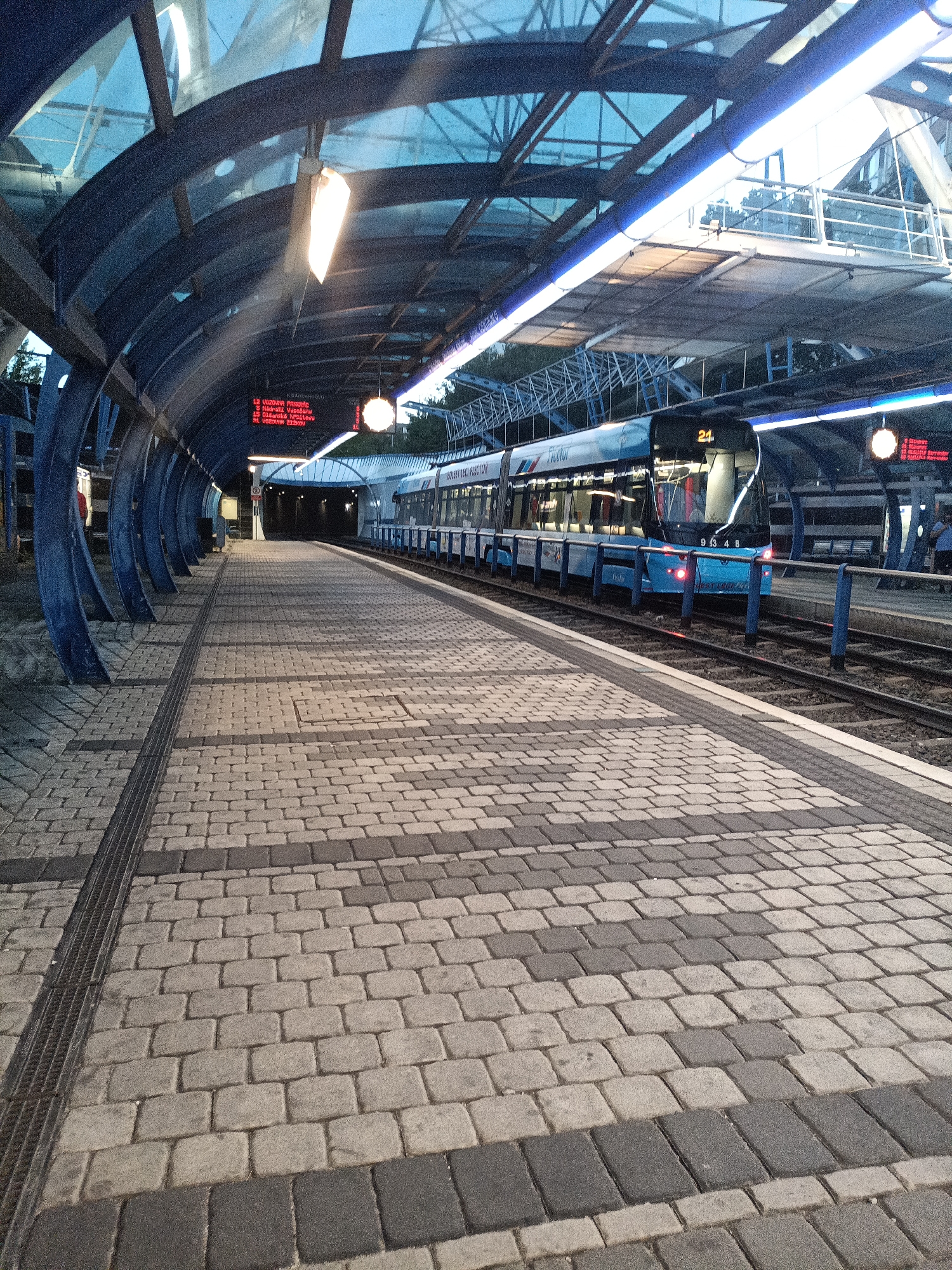 The image depicts a tram platform at a station in Prague, Czechia. A light blue tram, numbered 21, is stopped at the platform, its headlights illuminated. The platform itself is paved with gray and darker gray interlocking bricks, with a metal drainage grate running along the edge closest to the tracks. Above the platform is a modern, curved canopy constructed with blue metal supports and a translucent blue roof, which is illuminated by interior lights. Red digital signs displaying train information are visible above the entrance to the tunnel where the tracks disappear. The scene is captured in the evening or early morning light, with artificial lighting casting a blue hue over the environment. The atmosphere suggests a functional urban transport hub during off-peak hours.