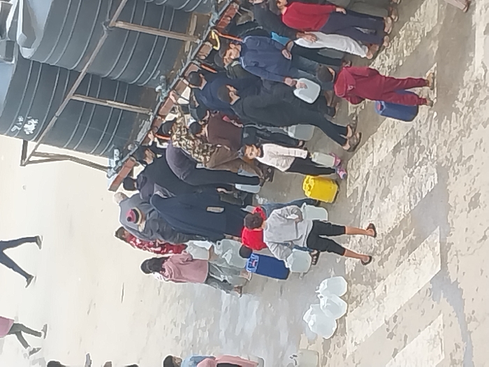 In the city of Khan Yunis, Palestinian Territory, an outdoor scene depicts approximately 20-30 individuals, comprising adults and children, gathered around a water distribution point. The distribution point consists of at least six large, black, horizontally ribbed cylindrical water tanks mounted on a metal support frame, positioned on the right side of the image. People are actively collecting water from spigots at the base of these tanks, bending over to fill various containers.
Numerous clear plastic jerrycans of differing sizes, along with several yellow and one blue jerrycan, are visible. Some containers are placed on the ground, while others are being carried. The paved ground, which features white painted stripes of a crosswalk, appears wet in various areas, suggesting water spillage. Individuals are dressed in casual clothing, including hoodies, jackets, and a dark blue hijab on one woman. A boy in a grey sweatshirt carries two clear jerrycans, a girl in a white sweatshirt carries one, and a boy in a burgundy outfit carries a blue jerrycan. The group is closely clustered around the tanks, indicating a communal gathering for water collection.