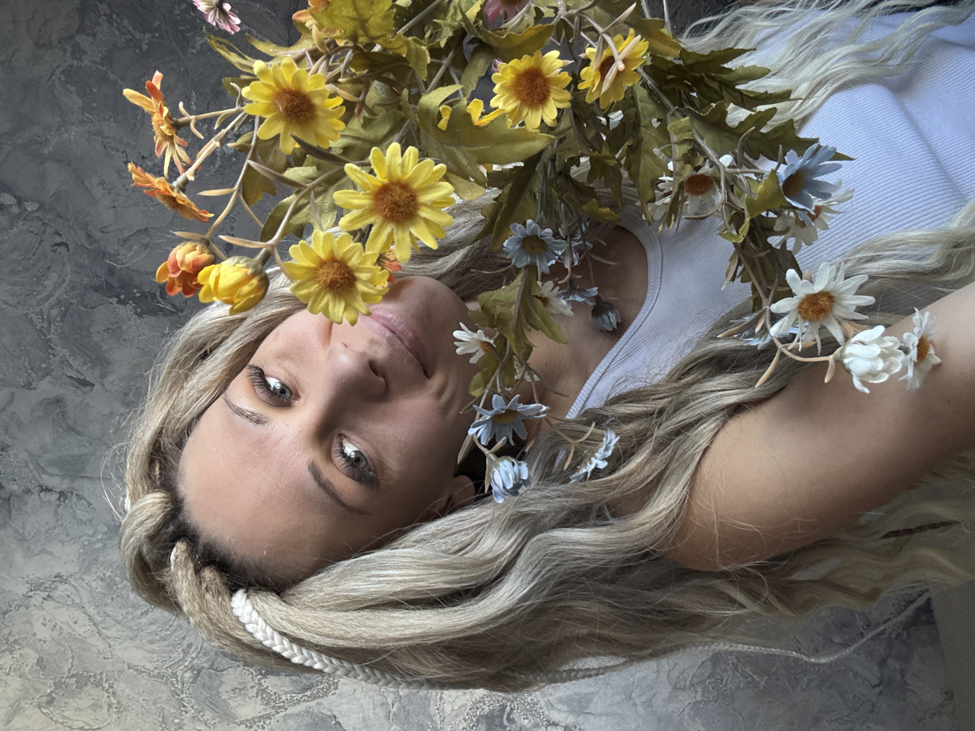 A woman lies on a gray surface, likely a floor or similar flat area with a mottled texture. She is positioned at an angle, facing the camera. She has long, blonde, braided hair, with some flowers woven into it and scattered around her. Her complexion is fair, and she appears to be wearing minimal makeup. She is wearing a white sleeveless top. Above her is a cluster of yellow and orange flowers, intermixed with smaller white and blue flowers. The specific location in Rusanovo, Russia, cannot be determined from the image.