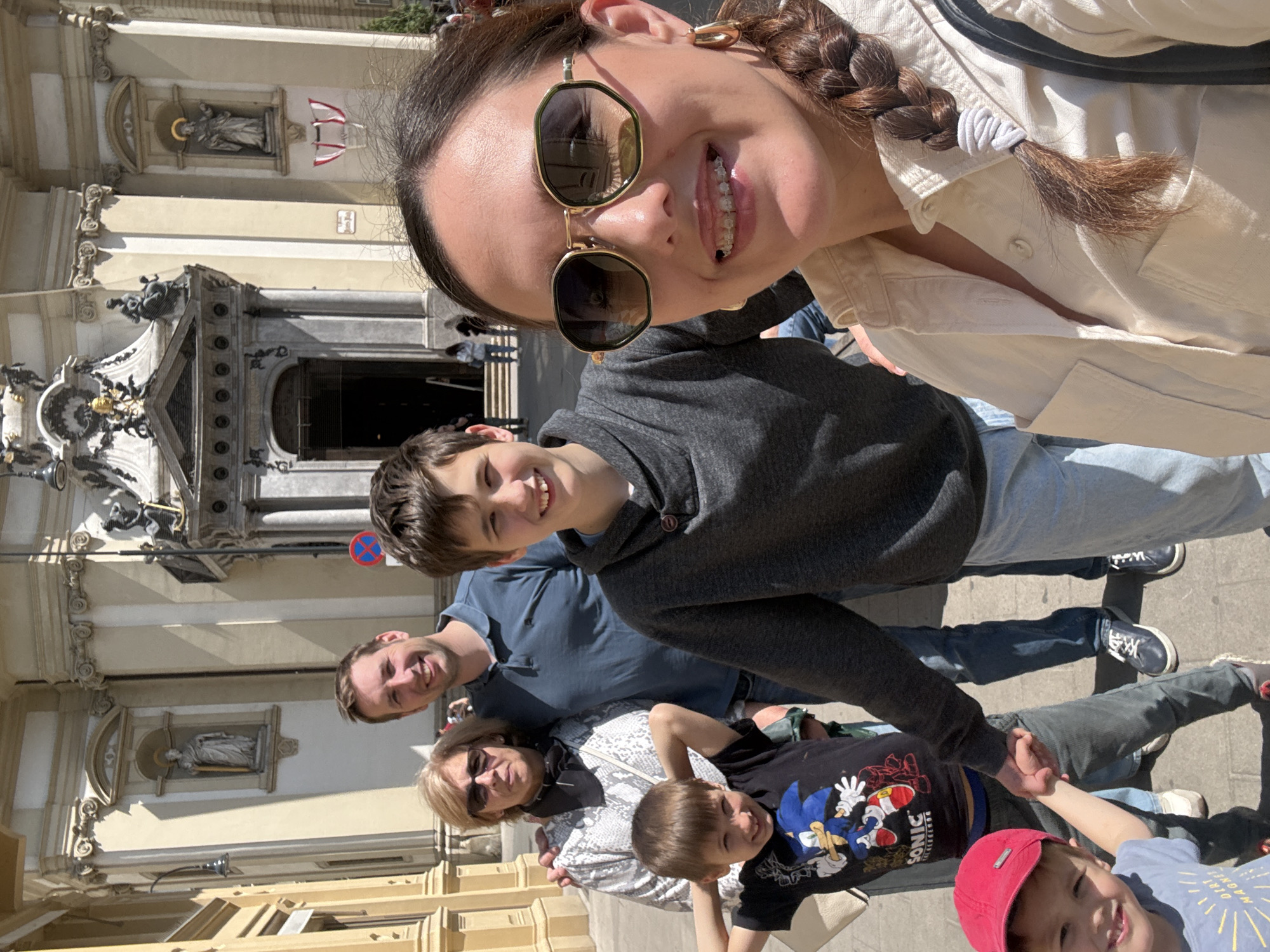The image shows a selfie of a group of people in Vienna, Austria. The foreground shows a woman with sunglasses and a braid. Behind her, a boy is smiling. Behind them are two adults and two more children holding hands. The background features the façade of a building with statues, likely a church or historical building, indicating that the group is in a tourist area of Vienna. A no parking sign can be seen behind the boy. The time of day appears to be daytime, likely during the day. The weather conditions seem sunny and warm.
