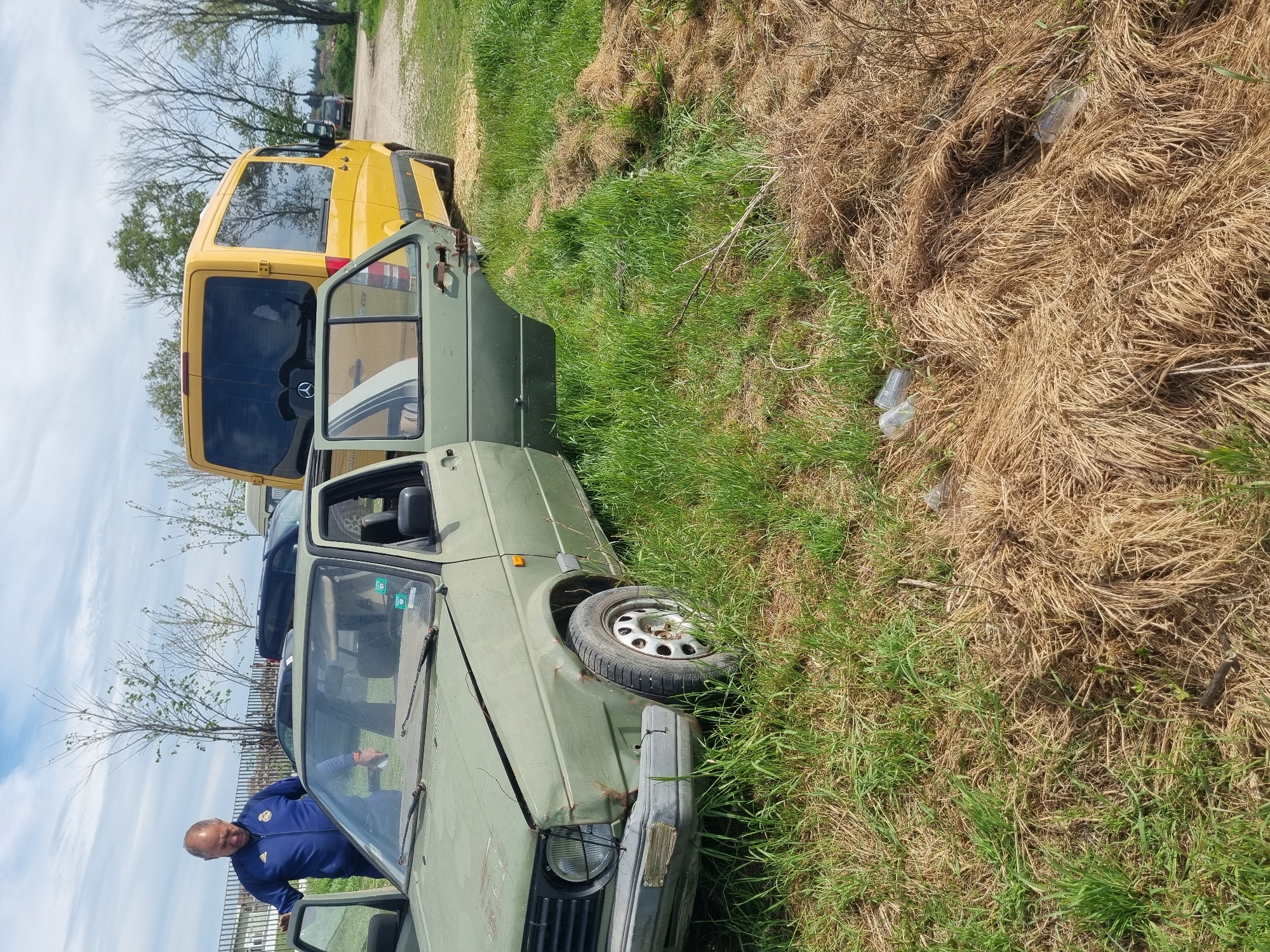 The scene is located in what appears to be a field in Ovcharovo, Bulgaria. An older man wearing a blue jacket is standing near the open door of a dilapidated, olive green car. A yellow Mercedes-Benz Sprinter van is parked nearby. The surrounding terrain consists of grass and dry, matted vegetation. There are also discarded plastic bottles visible on the ground. The condition of the green car suggests it has been abandoned or unused for some time.