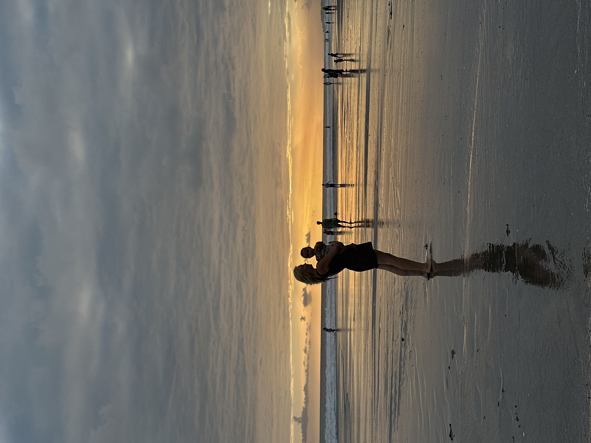 A silhouette of a woman holding a baby stands on a wet sand beach at sunset. The woman has blonde hair and is wearing a dark dress. The baby is facing away from the camera. In the background, the sun is setting over the ocean, casting an orange glow on the clouds and water. Other people can be seen walking on the beach in the distance. The sand is reflective, showing ripples and patterns from the receding tide. This scene is located on the beach in North Kuta, Indonesia.
