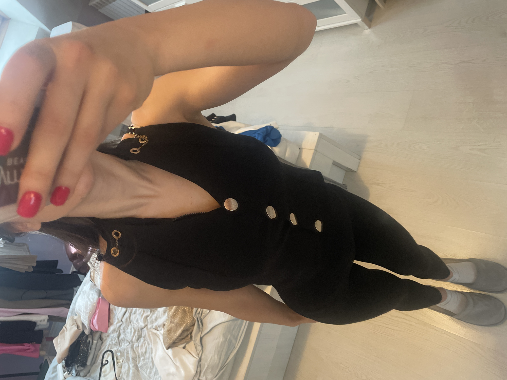 A person is taking a selfie indoors. They are wearing black pants with gold buttons down the side and grey slippers. The person's right hand is raised to hold the phone, revealing red nail polish. The background shows a bed with various items of clothing and storage furniture. The location is not identifiable as Sofia, Bulgaria based on the provided image.
