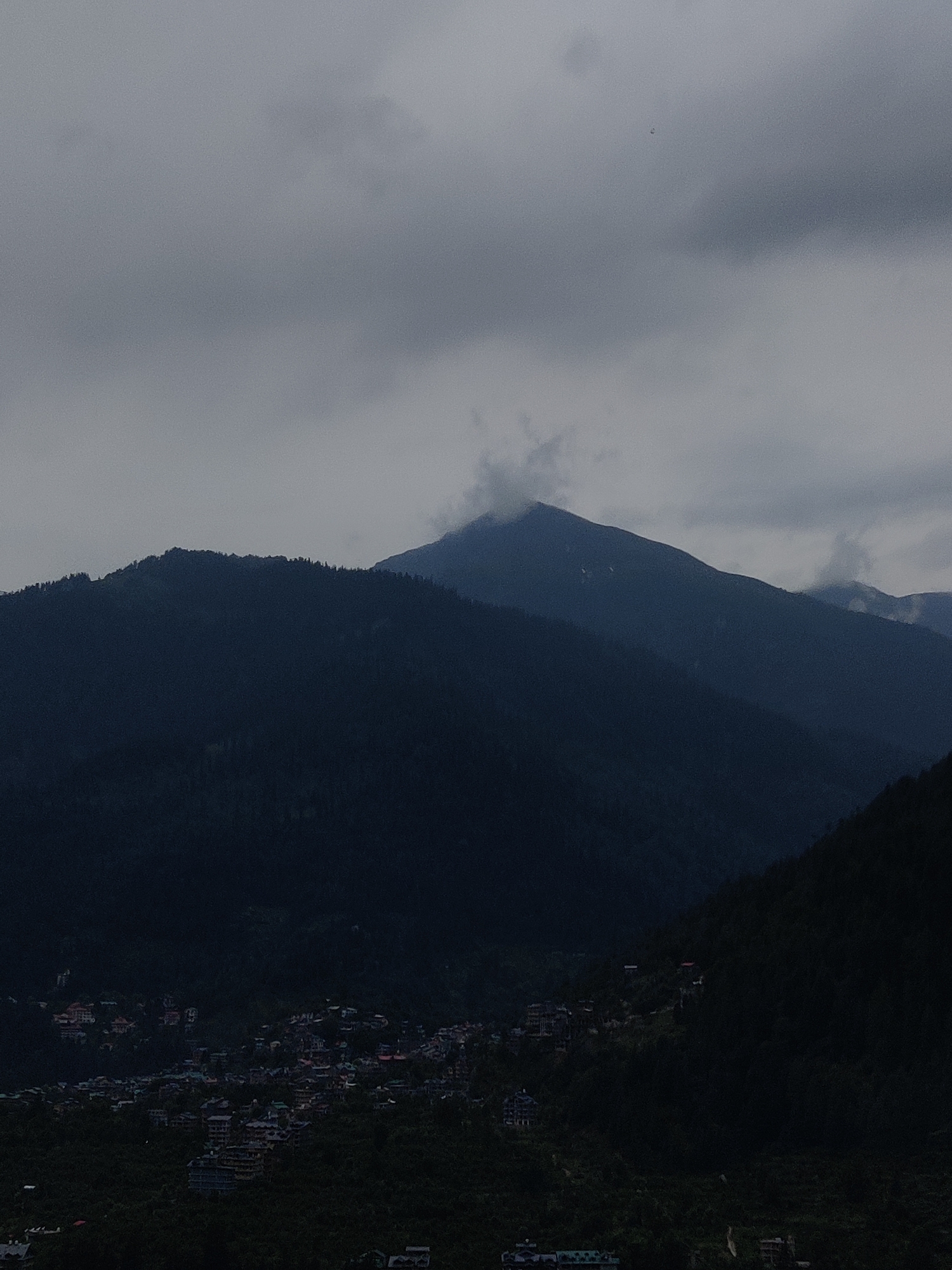 The image shows a panoramic view of a mountainous region. In the foreground, a densely packed village is nestled on the slopes of a lush green mountain. The houses, with their distinctive red-tiled roofs, are clustered together, giving the impression of a vibrant community.
Behind the village, the landscape rises into towering, dark green mountains, their peaks partially obscured by swirling clouds. The sky above is a moody gray, suggesting overcast or possibly rainy weather. The overall atmosphere is serene and somewhat mysterious, with the mist and clouds adding a sense of depth and grandeur to the scene.
The light in the image is subdued, indicating it might be early morning, late afternoon, or a heavily overcast day. The presence of buildings and the arrangement of the village suggest human habitation and activity within this natural setting. The location appears to be a hilly or mountainous area, likely in a region with a subtropical or temperate climate, given the dense vegetation. There are no visible signs of text in the image.