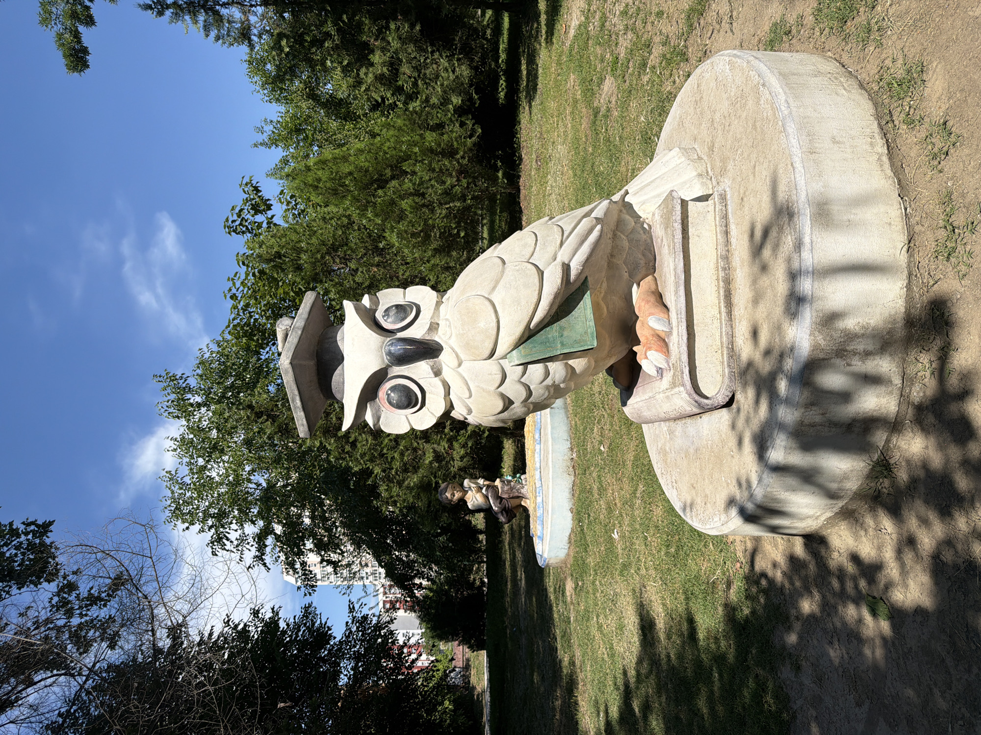 This is a medium shot, eye-level photograph taken outdoors on a sunny day in the city of Pazardzhik, Bulgaria. In the foreground, dominating the frame, is a light-colored, stone sculpture of an owl wearing a graduation cap. The owl is perched on top of an open book, with its talons resting on the pages. The owl has large, circular eyes with black pupils and a green cover visible under its wing, resembling a book or a folder.
In the mid-ground, to the left and slightly behind the owl sculpture, is another stone sculpture of a young boy sitting on a tree stump, holding something in his hands. This sculpture is within a circular, blue-rimmed basin.
The sculptures are situated on a grassy area with scattered patches of dirt and fallen leaves, particularly in the lower left and right portions of the frame. Sunlight casts dappled shadows across the ground. In the background, a line of lush green trees creates a natural backdrop, with a tall, multi-story building with a pinkish hue visible through the trees to the far left. The sky above is a clear, bright blue with a few wispy white clouds. The overall scene appears to be a public park or garden area, likely a space for art installations.