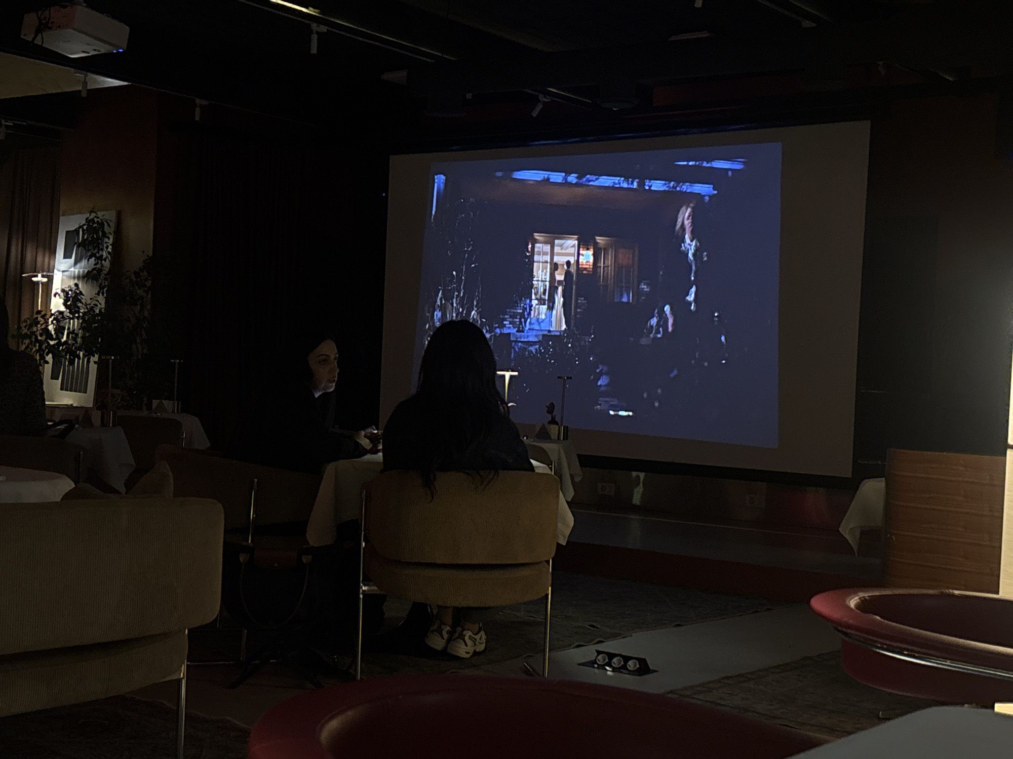 The image depicts an indoor setting, likely a lounge or restaurant, in Yerevan, Armenia. A large screen displays a video, potentially a film, in front of which people are seated. Two individuals are prominently visible: one, with dark hair, is seated with their back to the camera; the other is seated to the left, and is looking in the direction of the screen. The environment includes tables with white cloths, chairs with metallic legs and yellowish upholstery, and red furniture. The lighting is dim, and a projector is visible on the ceiling, casting light onto the screen.