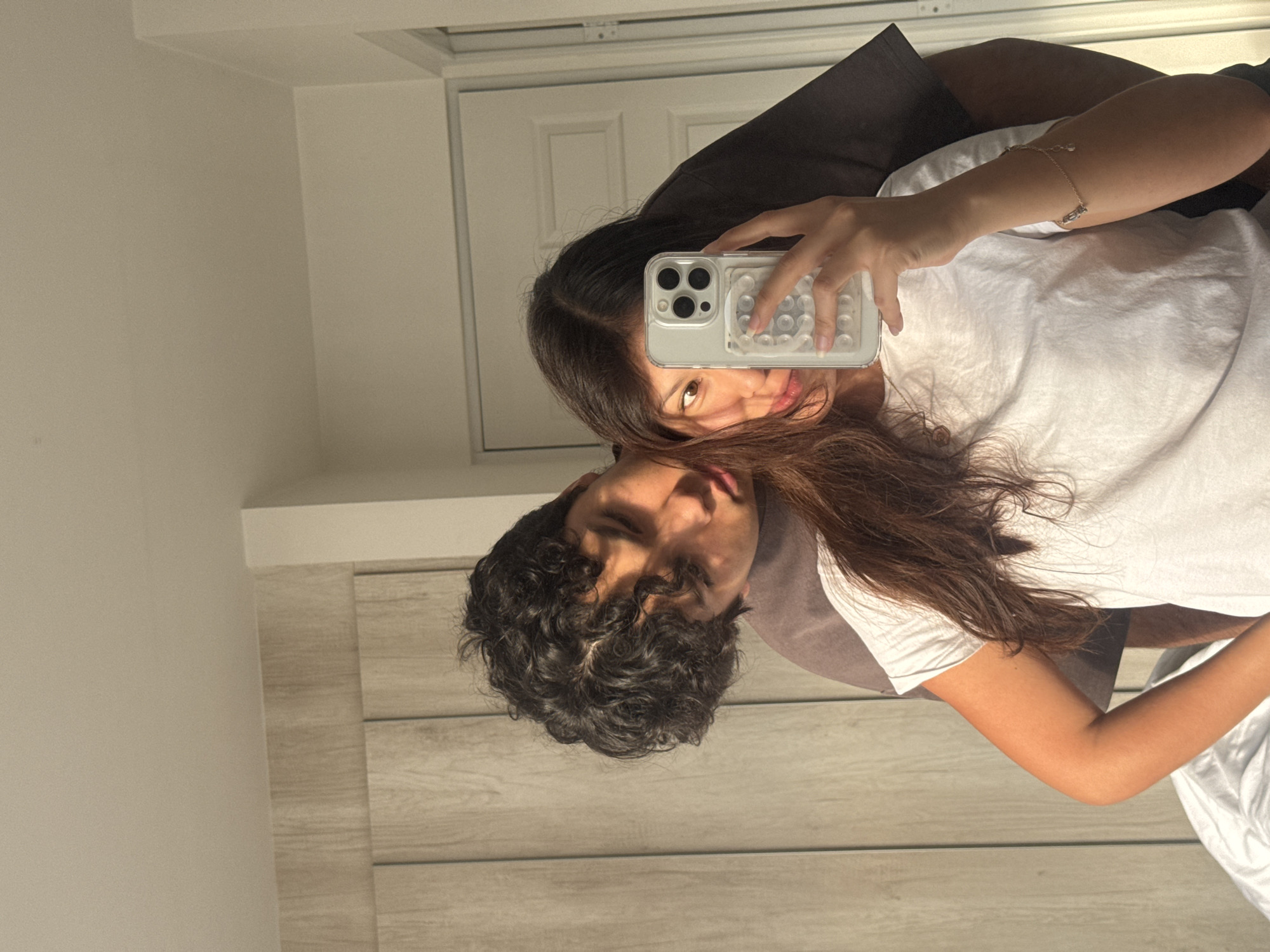The image shows a selfie taken in a mirror. A young woman with long brown hair is holding a smartphone with a clear case in her right hand, partially obscuring her face. Behind her, a young man with dark, curly hair is visible. Both appear to be wearing white shirts. The background includes a white door and a wooden-paneled wall. The location appears to be indoors, likely a residence. There is no explicit information connecting the image to San Francisco Ocotlán, Mexico.