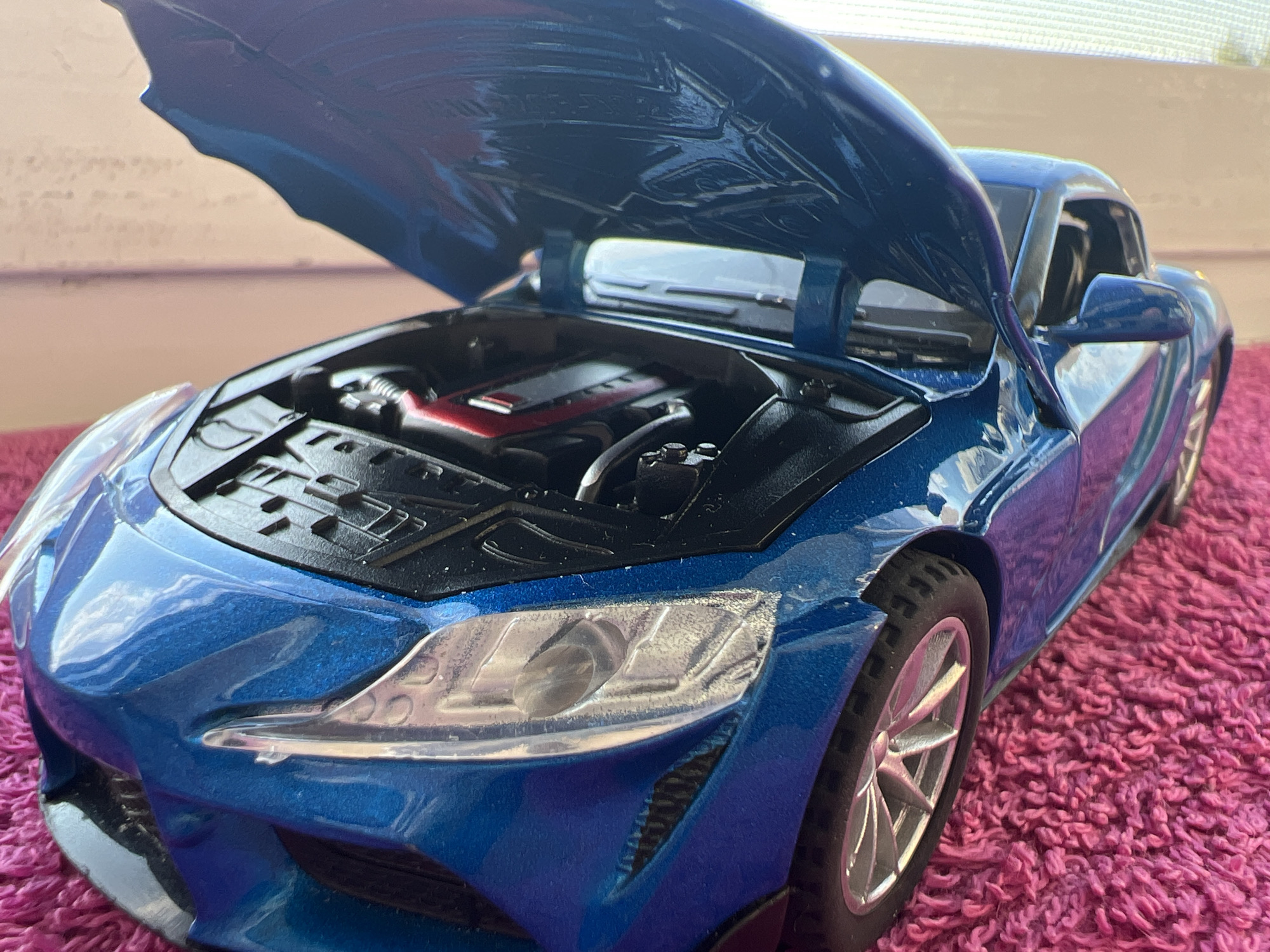 The image shows a close-up, eye-level, slightly angled shot of a blue toy sports car with its hood open. The car is positioned on a bright pink, fluffy surface, possibly a rug. The detail of the toy car is evident, with a visible engine compartment featuring red and black components. The car's headlights, side mirror, and wheel are also in sharp focus, showcasing its design. The background is blurred, suggesting it is out of focus or at a distance, with muted colors and a hint of a window or light source. There is no indication of the city of Omsk, Russia, in the image.