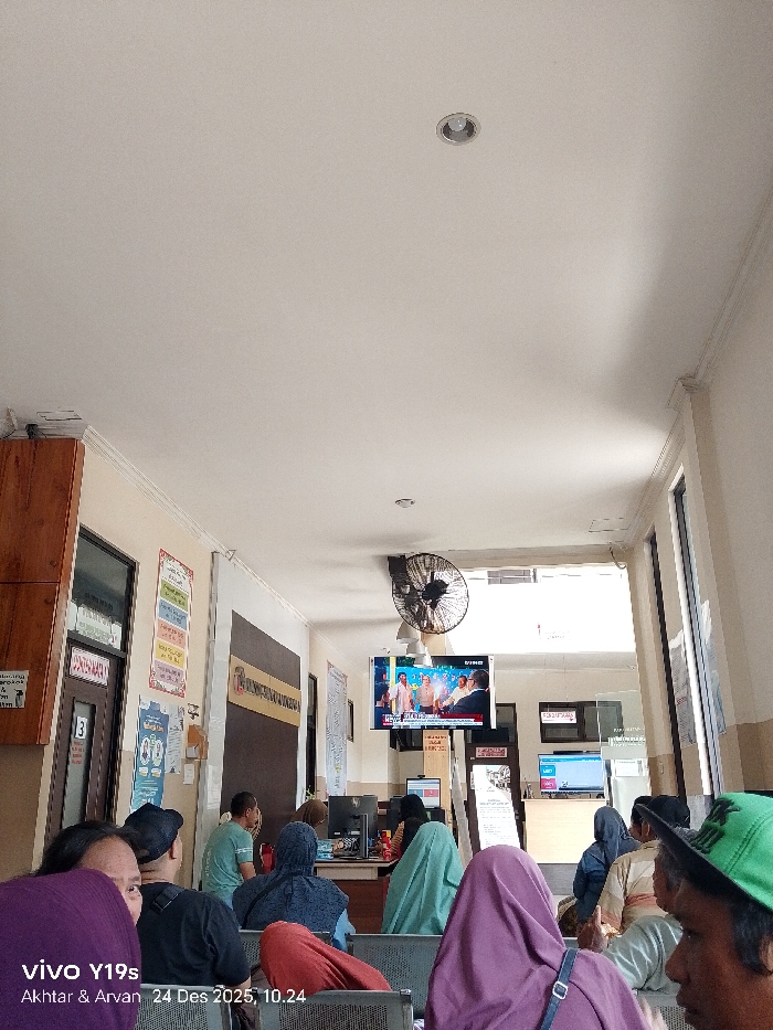 A waiting room is filled with seated people, some wearing head coverings. A wall-mounted television displays news. A ceiling fan rotates above the TV and a front desk area. Various signs and notices are posted on the walls. Light enters the room through windows at the far end.