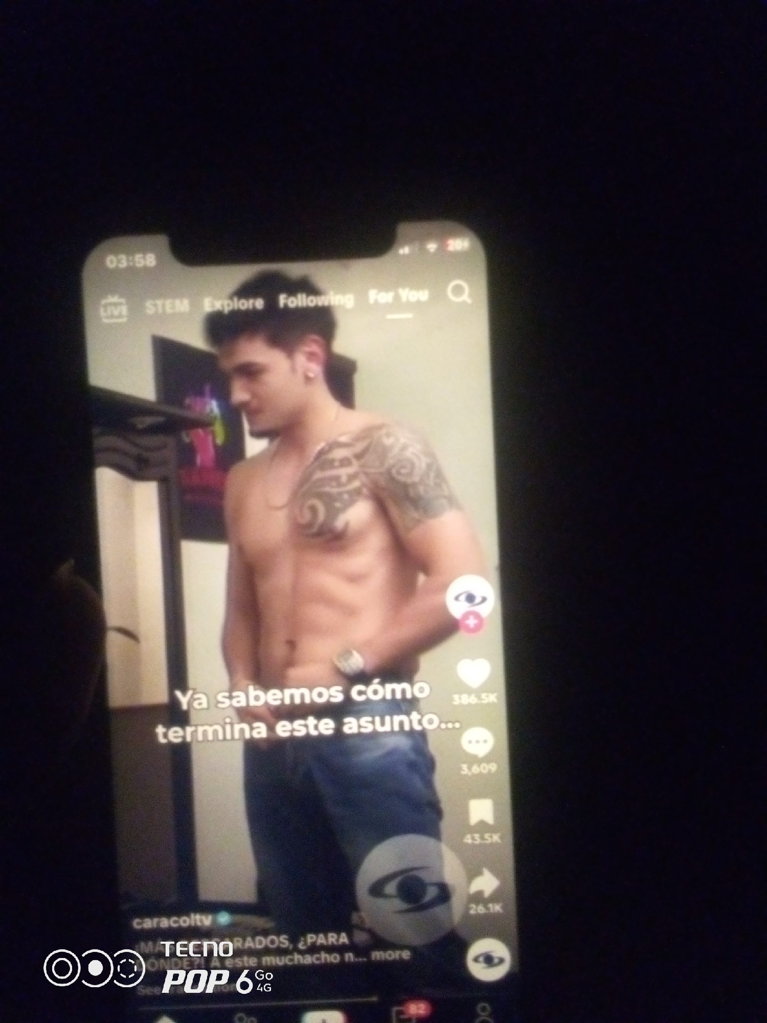 The image shows a close-up of a smartphone screen displaying a TikTok video. The video features a shirtless man with tattoos on his arm and chest. Text overlays on the video include "Ya sabemos cómo termina este asunto..." and a username "caracolty". Below the video, there are icons for likes, comments, shares, and other social media interactions, along with the text "TECNO POP 6 4G". The background of the video appears to be an indoor setting with some furniture visible. The phone is being held in a dark environment, likely at night. There is no information in the image to indicate any specific location in the city of Howayi, Nigeria.
