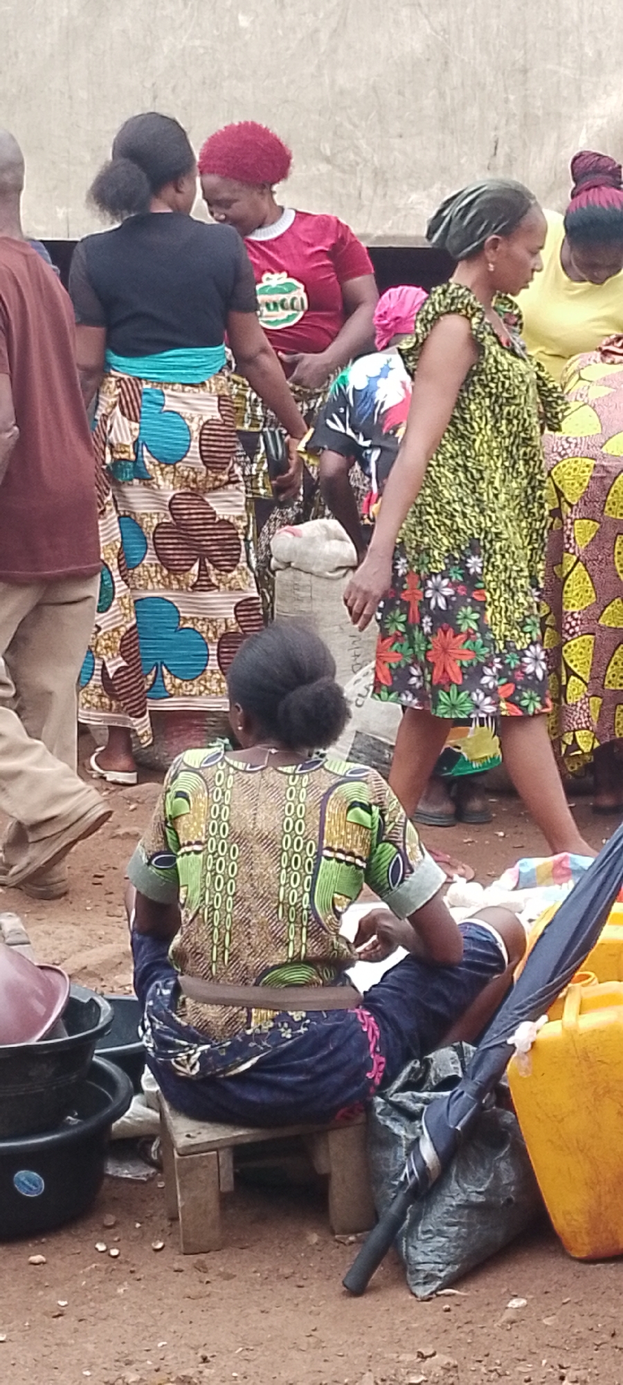 The image depicts an outdoor scene, likely a market, in Adikpo, Nigeria, with several individuals present. In the foreground, a woman is seated on a wooden stool, facing away from the camera, wearing a patterned brown and green top and a blue patterned skirt. Beside her are several black plastic containers and a large yellow jerrycan. To her right, a dark grey trash bag is visible, with a black umbrella leaning against it.
Behind the seated woman, other individuals are visible. One woman, wearing a green and yellow patterned dress with a floral hem, stands to her right, looking to the side. Further back, several other women are gathered. One is wearing a red t-shirt and another a black top with a teal sash. A woman in the background wears a yellow t-shirt. Many of the women are wearing brightly colored, patterned African print fabrics. The ground appears to be dirt or packed earth, consistent with an outdoor market environment. The background features a textured, light-colored wall. The overall atmosphere suggests a busy, communal gathering.