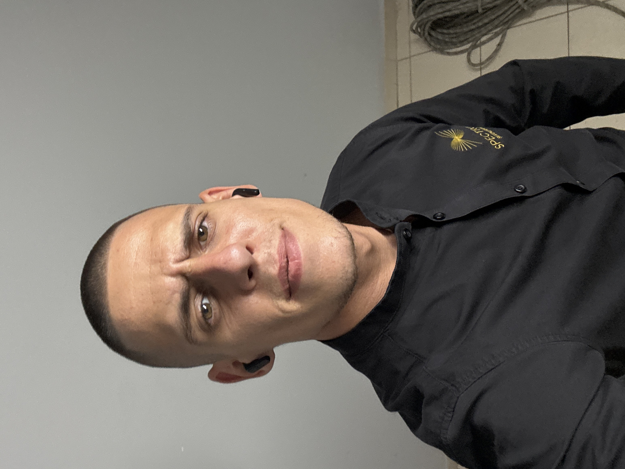 A man with a buzz cut and light brown eyes is looking directly at the camera with a furrowed brow and a slight smirk. He is wearing a black collared shirt with buttons down the front and a small, gold logo on the left side of the chest that reads "SPECTRA" with an abstract sunburst design above it. He has black wireless earbuds in both ears. The background is a plain, light gray wall. To the right of the man, a coil of thick, gray rope is visible against a tiled floor. The overall scene appears to be an indoor setting, possibly a workshop or storage area, with no identifiable landmarks to suggest a specific location within Sofia, Bulgaria.