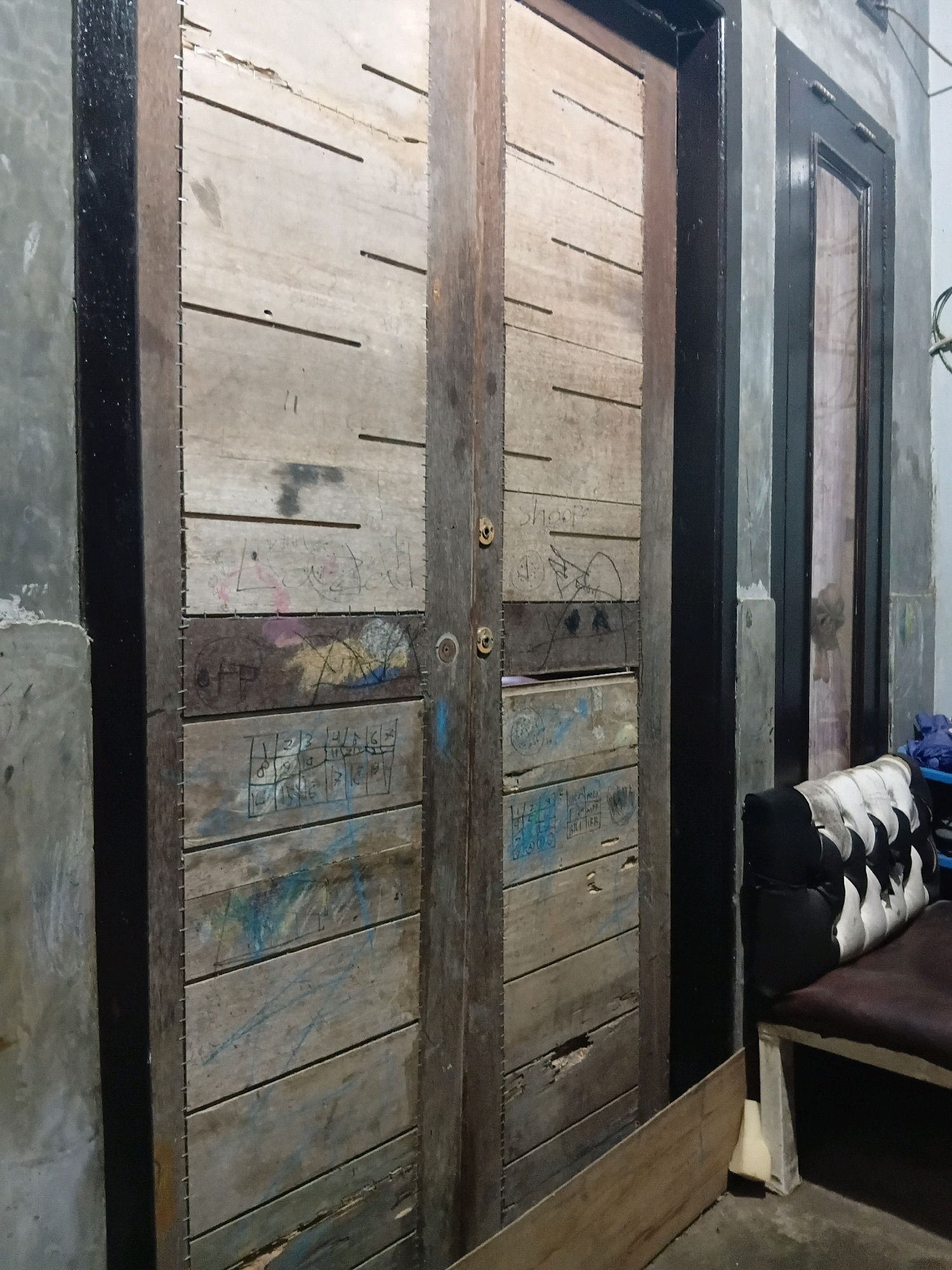 The image shows a close-up of a weathered wooden double door in Cikal, Indonesia. The doors are made of horizontal planks of wood, some of which are cracked and damaged. They are secured with metal staples along the vertical seams. Both doors have graffiti on them, including what appears to be numbers, symbols, and scribbled text. There are also two circular metal knobs on the right door, likely door handles. To the right of the wooden doors, there is a dark-framed door with glass, and next to that, a segment of a concrete wall. In the lower right corner, a cushioned bench with a black and white patterned seating cushion rests on a dark brown base, which is supported by light-colored legs. A light brown wooden plank is propped up against the right side of the double doors. The overall environment appears to be dimly lit and possibly an outdoor or semi-outdoor space.