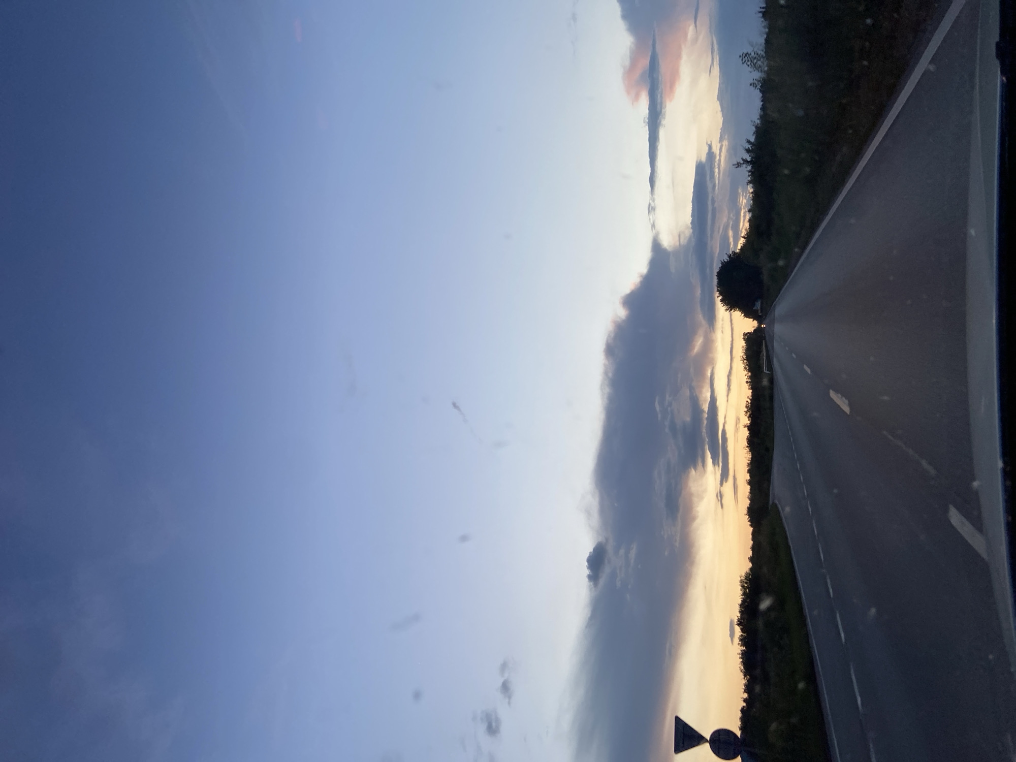 The image shows a view from a vehicle, driving on an asphalt road towards the horizon at dusk or dawn. The road has intermittent white lines. The right side of the road is lined with green vegetation. The sky occupies a significant portion of the image, displaying a gradient of colors from blue to shades of orange and gray, indicating either sunrise or sunset. There are scattered clouds visible in the sky. Road signs are seen to the lower left of the frame. The location is near Dragijevo, Bulgaria.