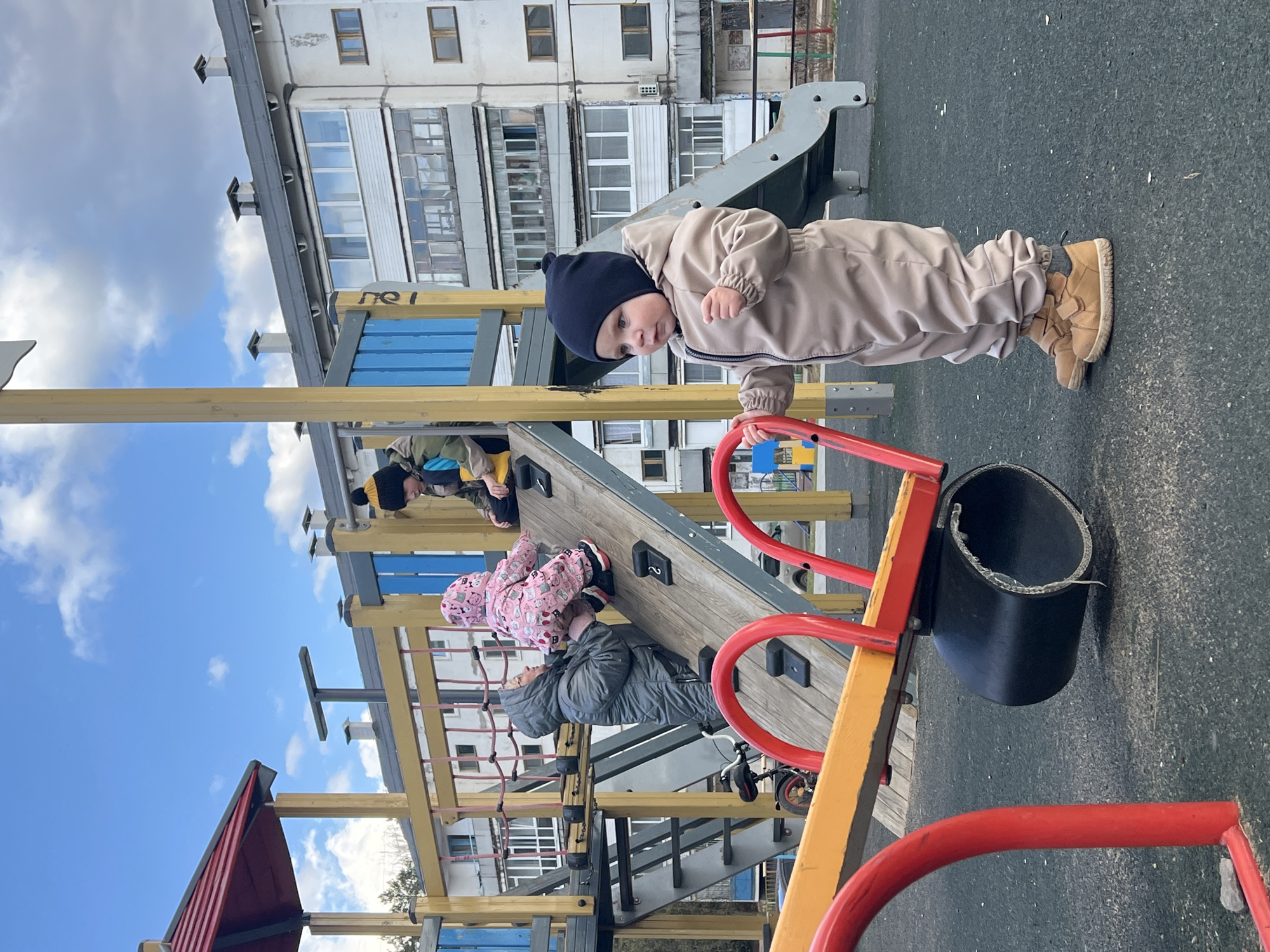 This media file captures an active scene at an outdoor playground, likely in Noviy, Russia. The image is composed with a slightly upward angle, showcasing a multi-story residential building against a partially cloudy blue sky in the background.
The primary subject is a young child, appearing to be a toddler, dressed in a warm, practical beige jumpsuit and a dark blue beanie. This child stands upright on the dark playground surface, holding onto a red metal bar of the play structure. They are looking directly at the camera with a curious expression. Their light brown boots are clearly visible.
Around and behind the central child, several other individuals are engaged in play on a wooden and metal playground structure. One child, wearing a pink patterned suit and a hat, is actively climbing a wooden wall fitted with black climbing holds, some marked with the number '2'. Below this child, another person in a grey padded jacket is also on the climbing apparatus. The upper body of a third person, in a yellow and blue jacket with a patterned hat, can be seen further up the structure.
The playground equipment includes a wooden climbing wall, yellow wooden beams, blue panels, red metal railings, and a grey slide partially visible in the background. A black bucket-like object is attached to a red bar. The ground beneath the play area is a dark, possibly rubberized, safety surface. The residential building in the background features multiple windows and enclosed balconies, characteristic of urban or suburban environments. Faintly visible on a blue panel of the play structure are the Cyrillic letters "пе l". The scene takes place during the daytime, likely on a cool day given the warm attire of the children.