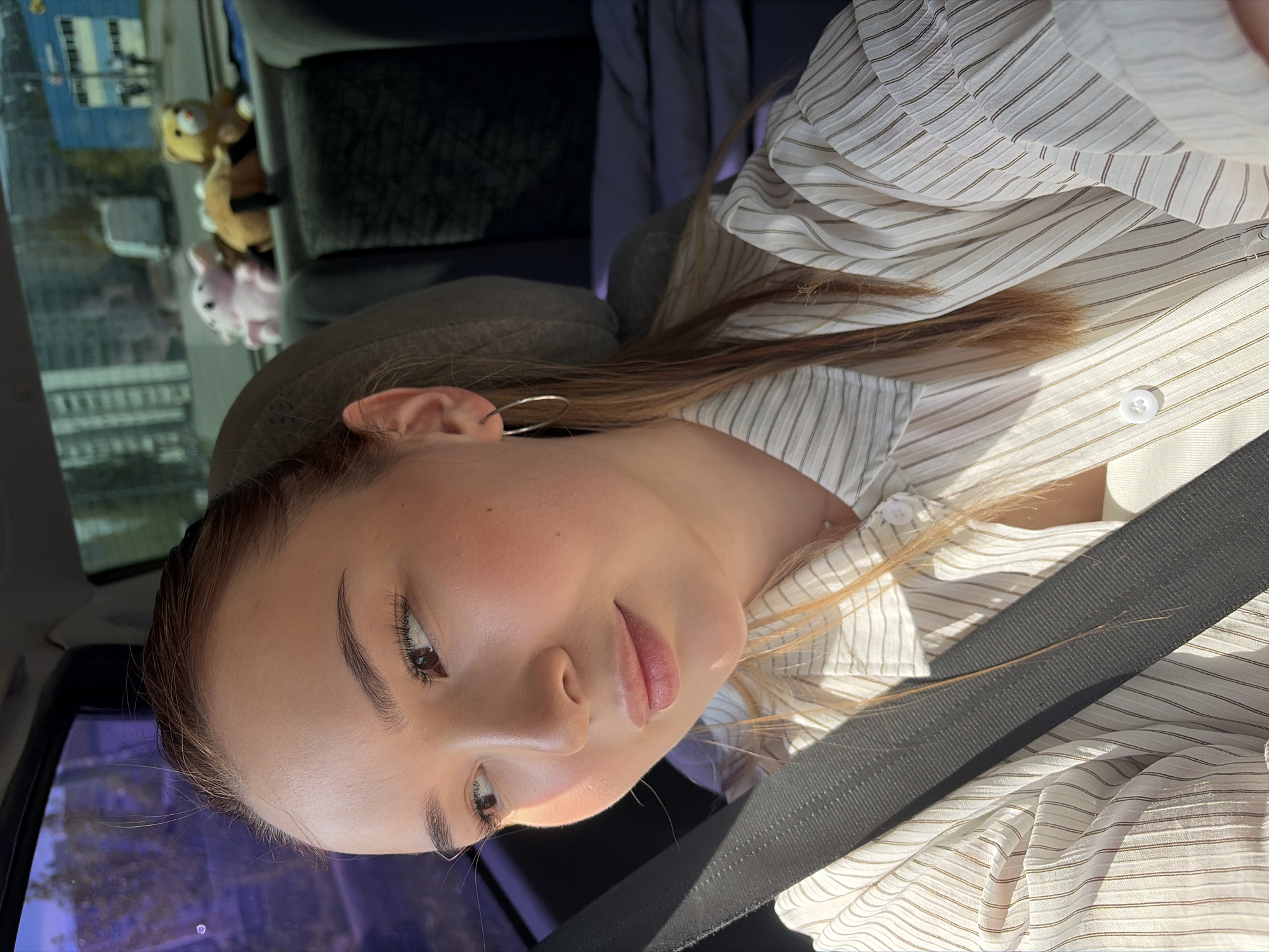 A young woman is seated in a car, secured by a seatbelt. She has fair skin, brown hair, and is wearing a striped, light-colored shirt. Outside the car window, buildings are visible, indicating an urban environment. Stuffed animals are visible in the rear of the car. The location is Chaikovsky, Russia.