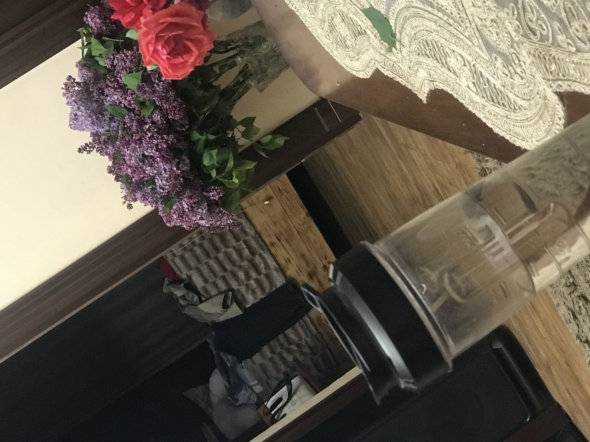 A high-angle shot captures a tabletop scene. The table is set with a lace runner. To the left of the runner, there is a bouquet of lilac and roses. Adjacent to the bouquet, a clear plastic blender is positioned on the table. Below the table is a cabinet, where the interior holds miscellaneous items. There is no evidence in the image to indicate that it is from Yerevan, Armenia.