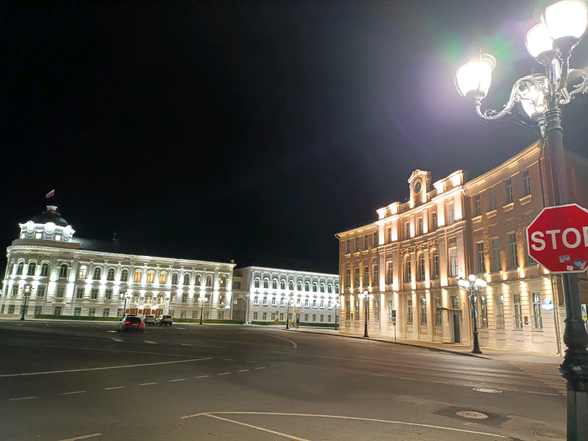 The image captures a nighttime view of the historic Imperial Traveling Palace in Tver, Russia, situated on a wide city square. The palace, a grand, neoclassical building, is illuminated, showcasing its ornate facade and domed roof topped with a Russian flag. Adjacent to it, another similarly styled building, likely part of the palace complex, is also lit. In the foreground, a street with visible lane markings curves around the square. Several cars are present, with one dark sedan in motion. A decorative street lamp with multiple lights stands prominently on the right side of the image, casting a bright glow. A red octagonal stop sign is mounted on a pole near the street lamp, indicating a traffic control point. The sky is dark, suggesting late evening or nighttime. The scene appears tranquil and devoid of significant human activity beyond the drivers of the vehicles.