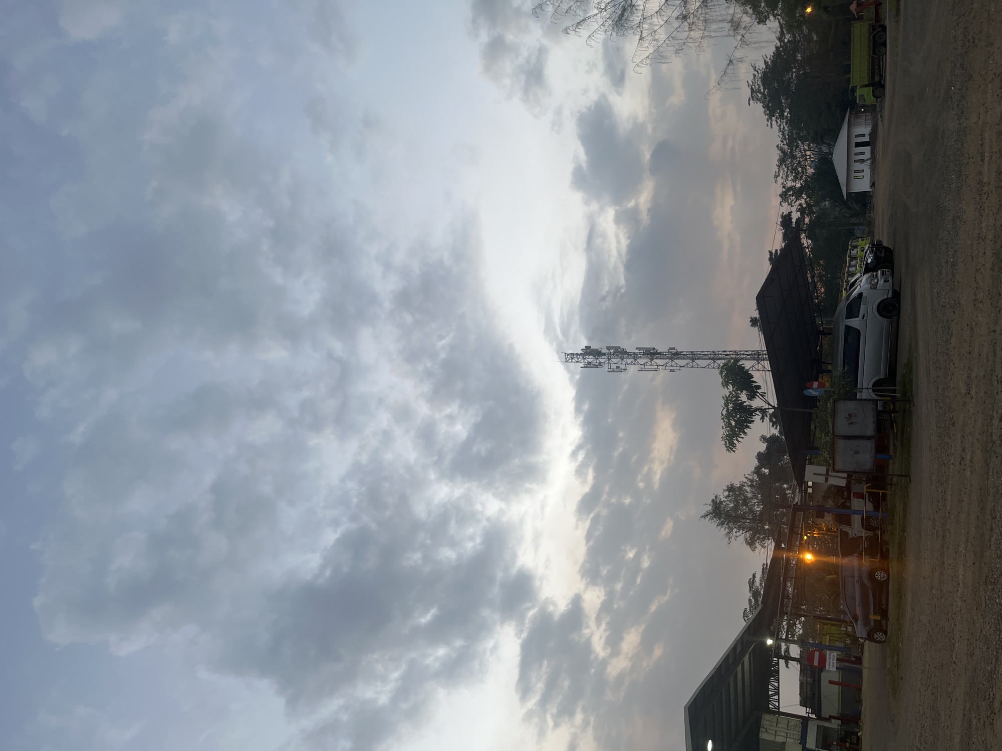 The image depicts an outdoor scene at dusk, possibly a truck stop or industrial area, with a cell tower prominently visible in the middle ground. The sky is filled with dramatic, overcast clouds, suggesting an impending change in weather or the end of the day. In the foreground, a dirt or gravel lot is visible, with several vehicles parked, including a dark-colored SUV, a silver van, and what appears to be a green truck. Structures with roofs, likely shelters or small buildings, are present in the left and center of the image, illuminated by overhead lights. Trees and vegetation frame the scene, with a few distant houses visible on the right. The overall atmosphere is subdued and somewhat industrial, bathed in the soft, diffused light of twilight.