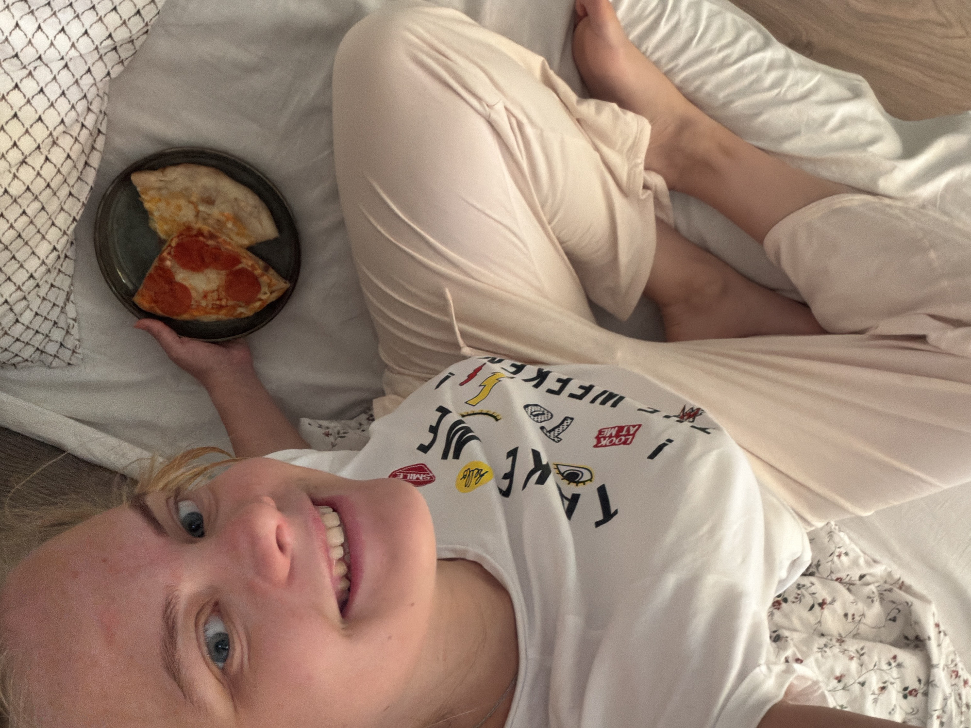 A woman lies on a bed in what appears to be a home environment. She is fair-skinned with blonde hair and blue eyes, smiling at the camera. She is wearing a white t-shirt with graphic designs. A plate containing two slices of pizza, one with pepperoni, rests in her hand. She is also wearing light pink pajama pants. The bed is covered in white bedding, including a pillow with a small floral pattern and another with a geometric design. The location is not identifiable as being in Chaikovsky, Russia, based solely on the visual information available in the image.