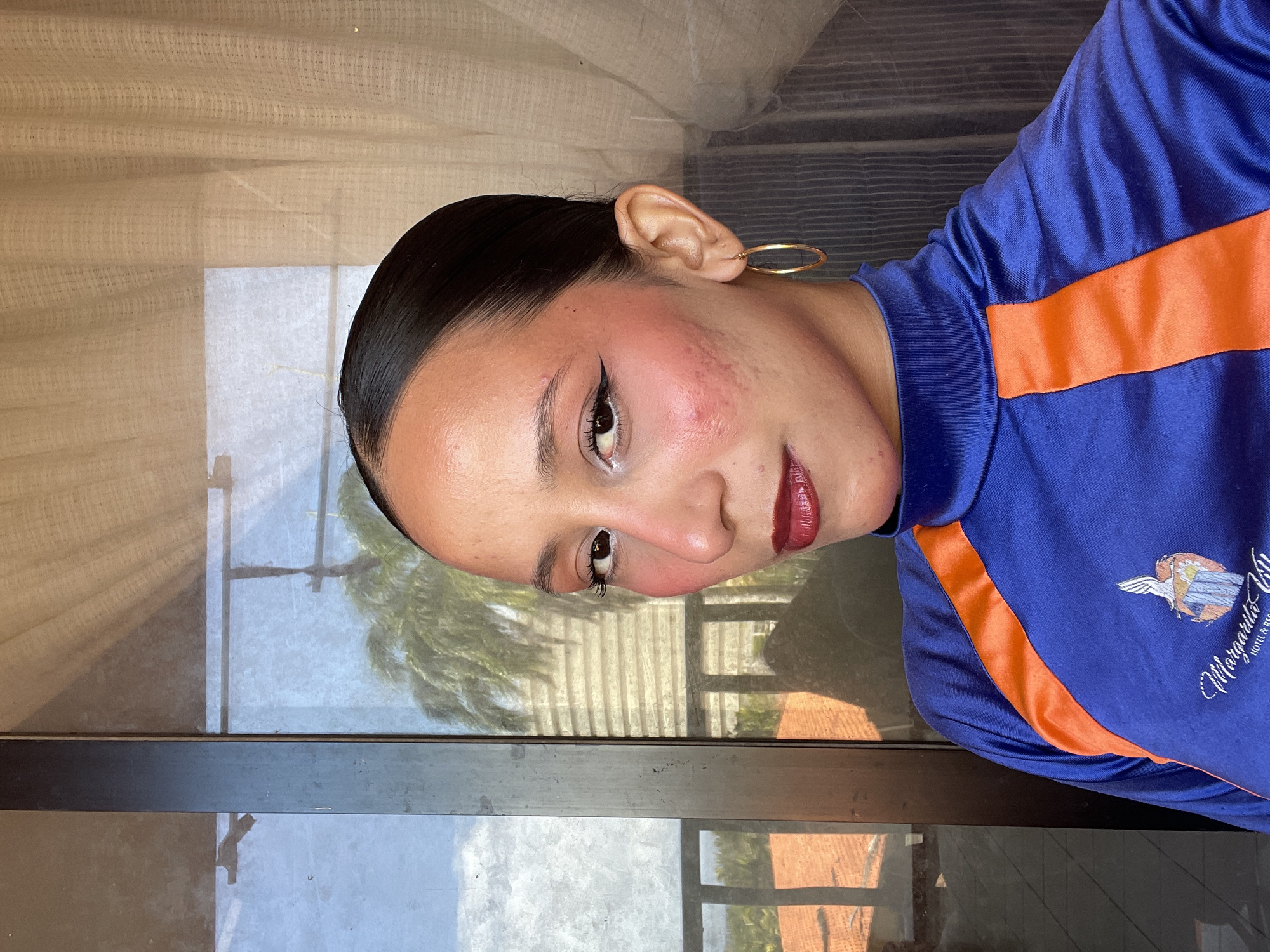 The image shows a close-up of a woman's face and upper torso, taken from a slightly low angle. She has dark hair pulled back, prominent cheekbones with blush, dark eyeliner, and dark red lipstick. She is wearing a blue and orange long-sleeved top with a high neckline, which features a logo on the left side of her chest. The logo is embroidered and appears to depict a pelican landing on a structure with water and palm trees. A gold hoop earring is visible in her right ear. The background is slightly out of focus, showing a window or glass door with reflections of palm trees, buildings, and a sky with clouds. The interior background consists of a light-colored, textured curtain. The overall lighting suggests it is daytime. While the specific location within Porlamar, Venezuela is not explicitly visible in the image, the presence of palm trees and reflections of buildings is consistent with a tropical or subtropical environment often found in coastal areas of Venezuela, and the clothing logo "Margarita U..." suggests a connection to Margarita Island, where Porlamar is located.